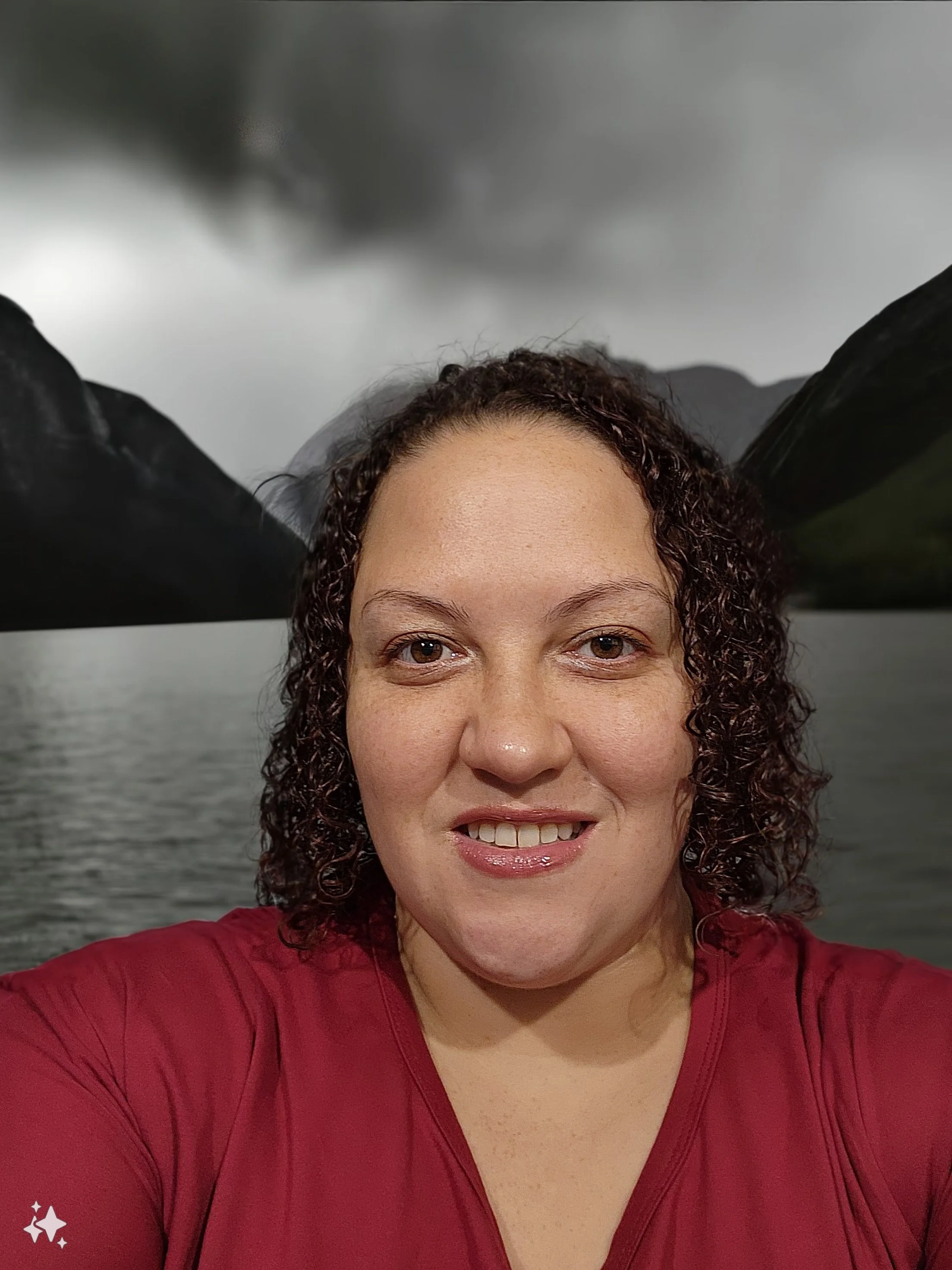 A woman smiling with shoulder-length curly hair, wearing a red top, in front of a body of water with mountains and an overcast sky in the background.