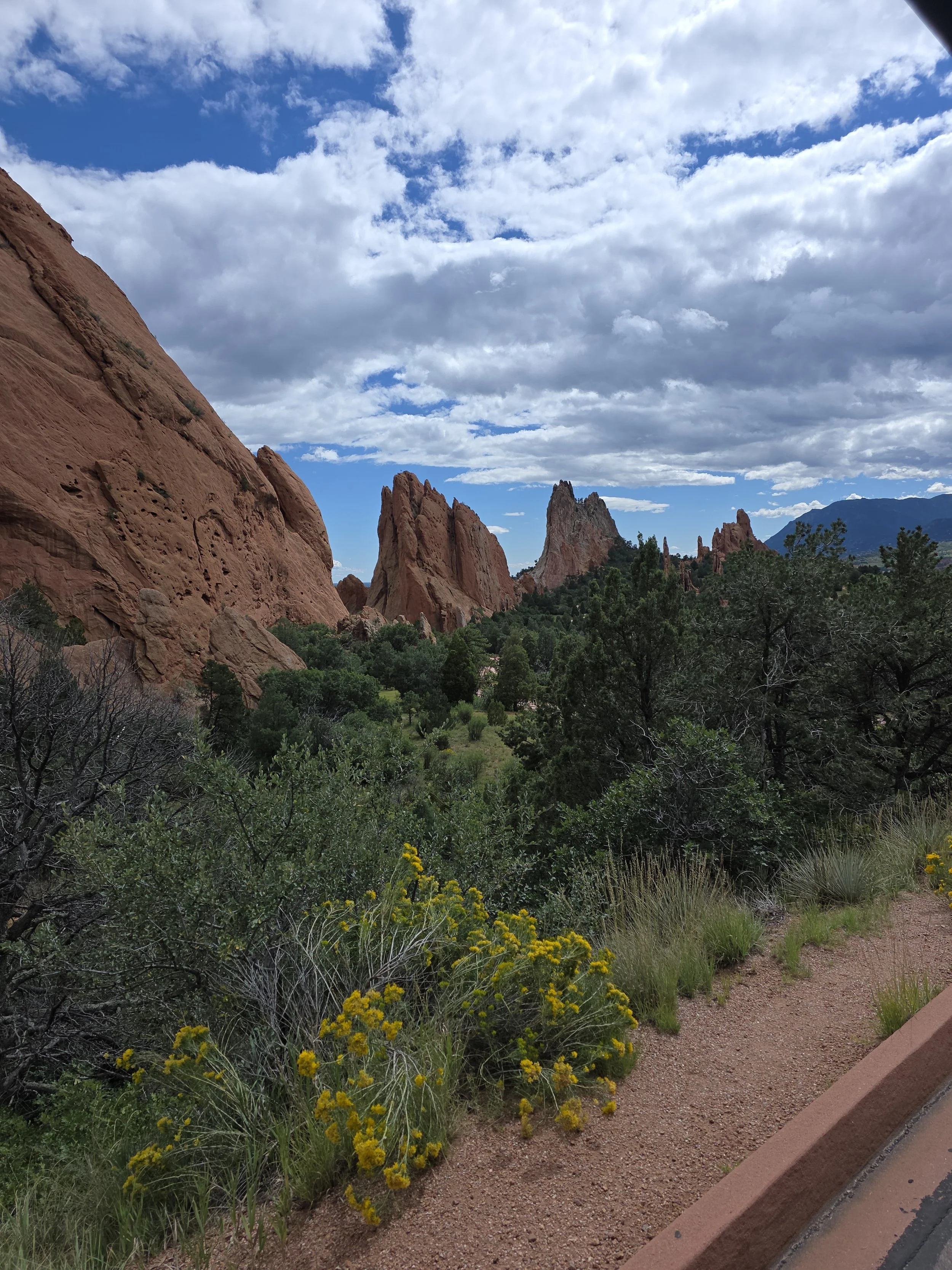 A scenic view of red rock formations with greenery and trees in the foreground, under a partly cloudy sky.