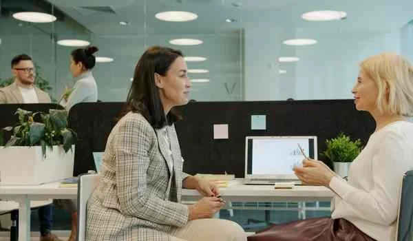 Two women sit facing each other at adjoining desks in a bright, modern open-plan office. One is wearing a grey checked blazer and the other a cream top. A laptop sits between them, with sticky notes on the partition behind them.