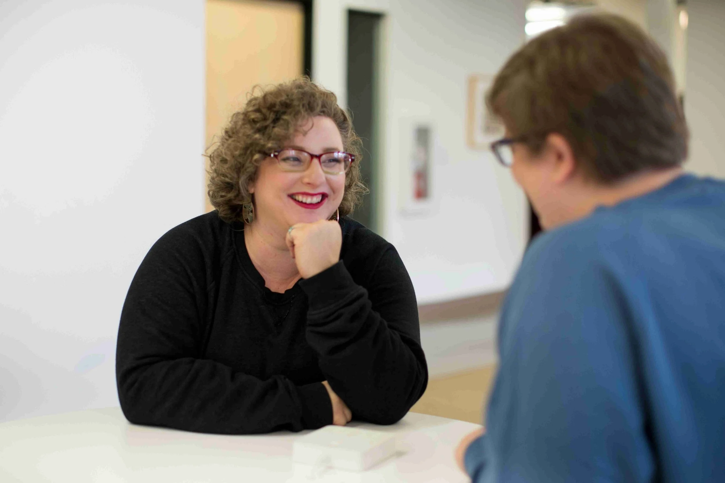 Two colleagues in conversation at a desk, one of them is listening and smiling. She is resting her chin on her hand. The other is out of focus with their back to the camera.