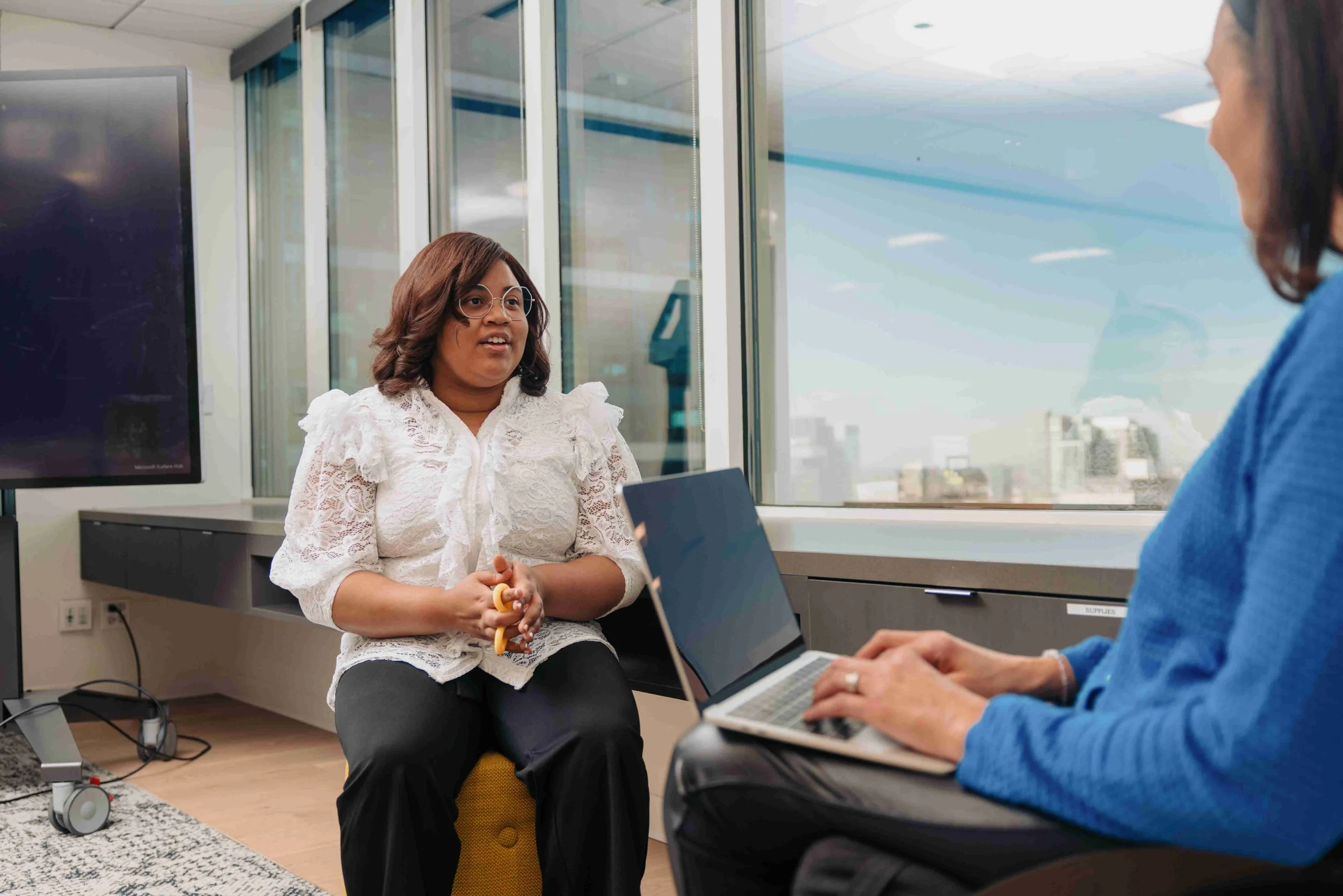 A woman sits in an office during a meeting with her manager. She holds a fidget toy in her hands while her manager types on a laptop.