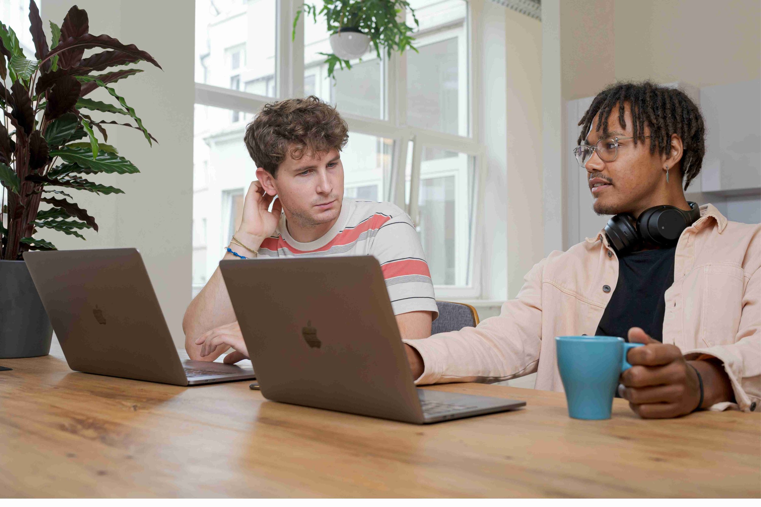 Two male colleagues sit side by side at a table in front of their laptops. One is holding a cup of coffee and is in conversation as his colleague looks over at his screen and looks pensive.