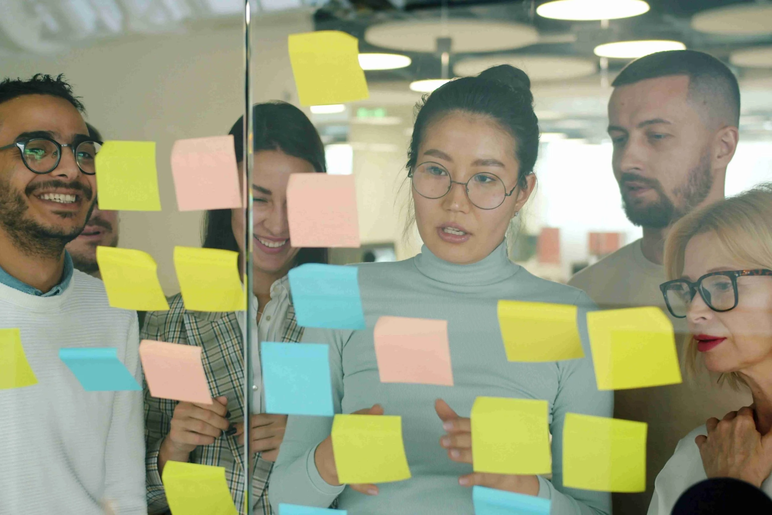 A small group of colleagues in a planning session, making conversation and reference to sticky notes stuck on the glass wall.