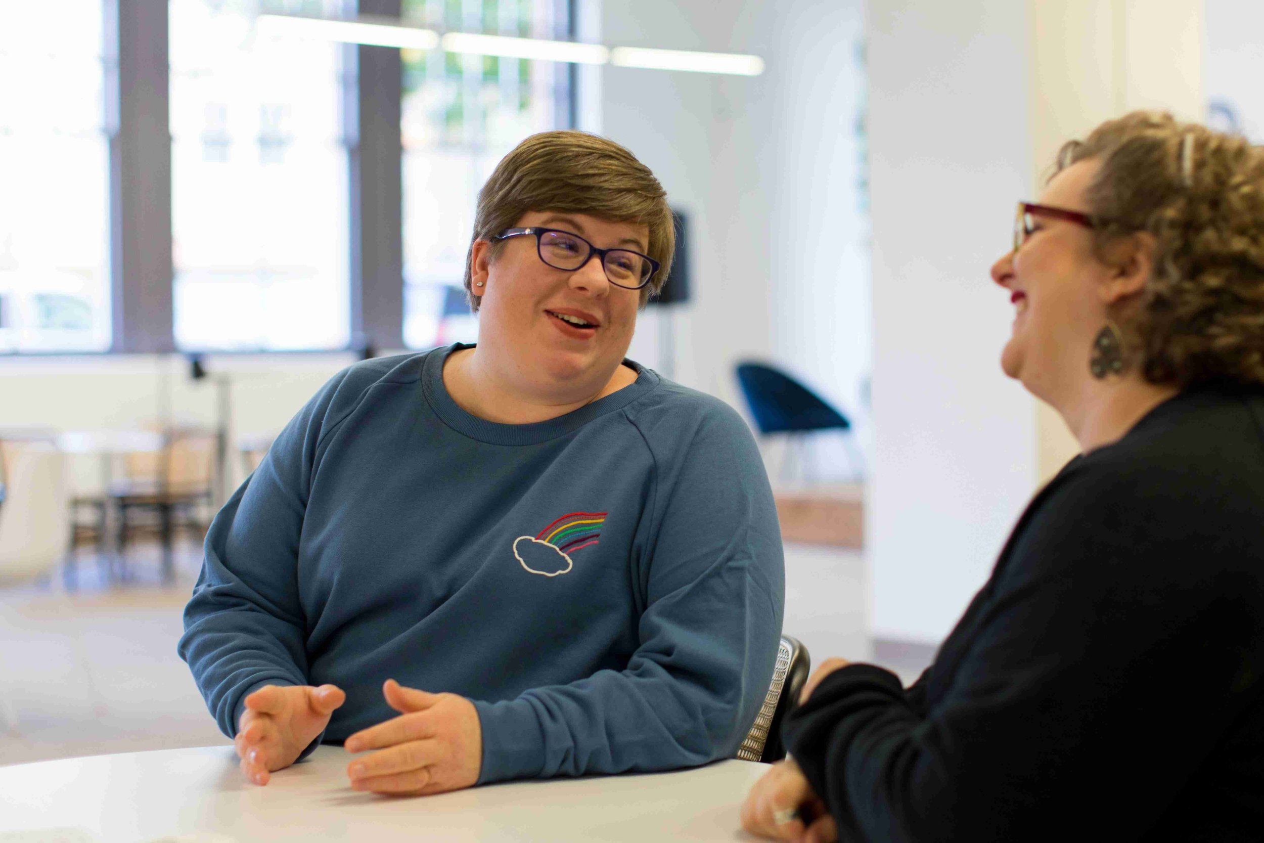 Two people smiling at each other sitting at a desk having an interview. The office is otherwise empty and the person being interviewed is comfortably dressed in a rainbow embroidered sweatshirt and appears relaxed