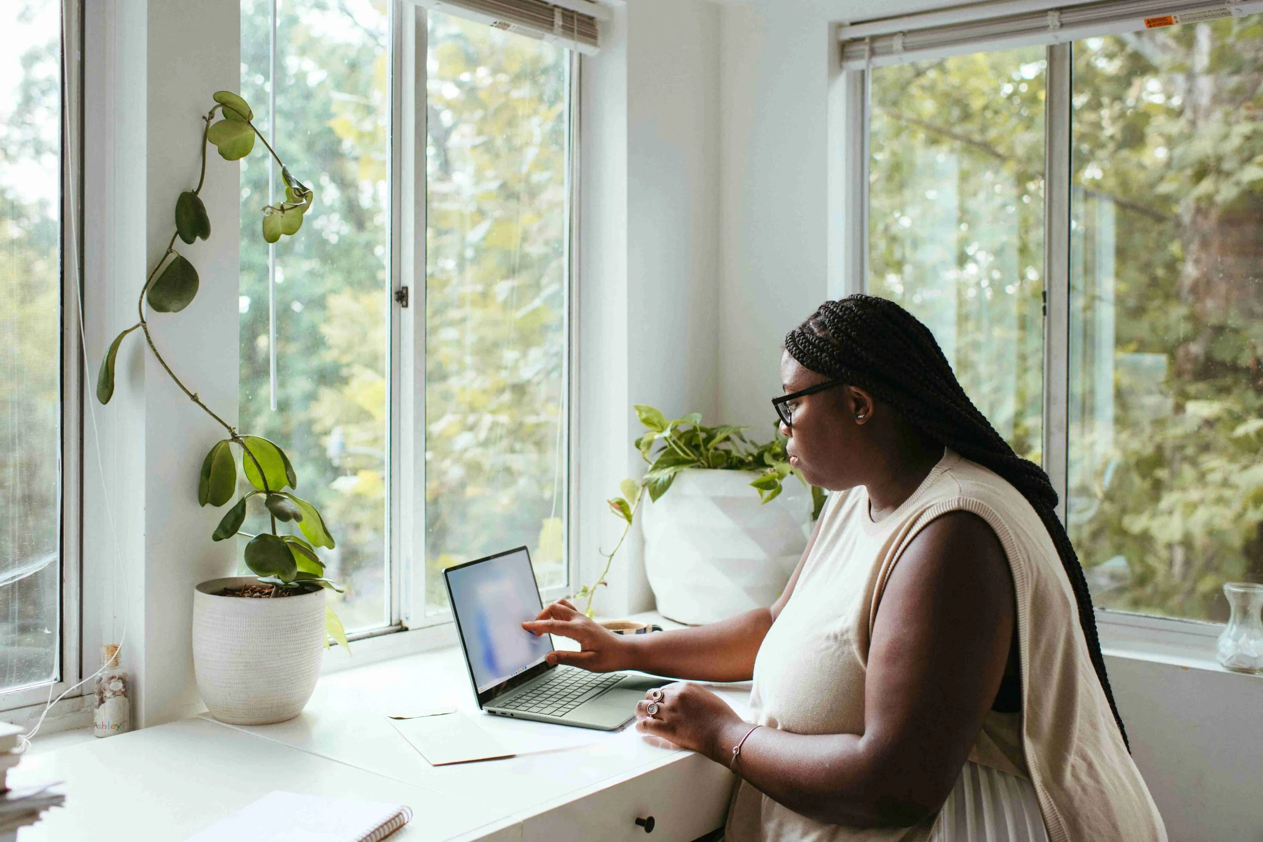 A woman sits in a light-filled window surrounded by plants. She is working on her laptop as she reviews the contents of her screen, blurred to the viewer.