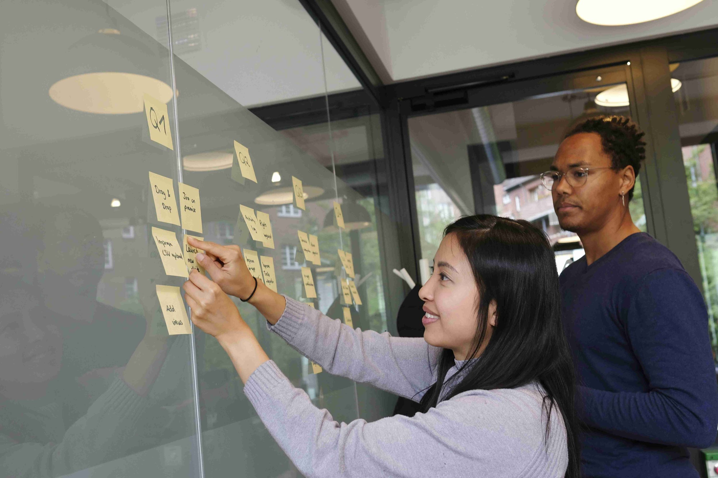 Two colleagues in front of a wall with sticky notes attached to map out a process. One of them is in action sticking another to the existing notes.