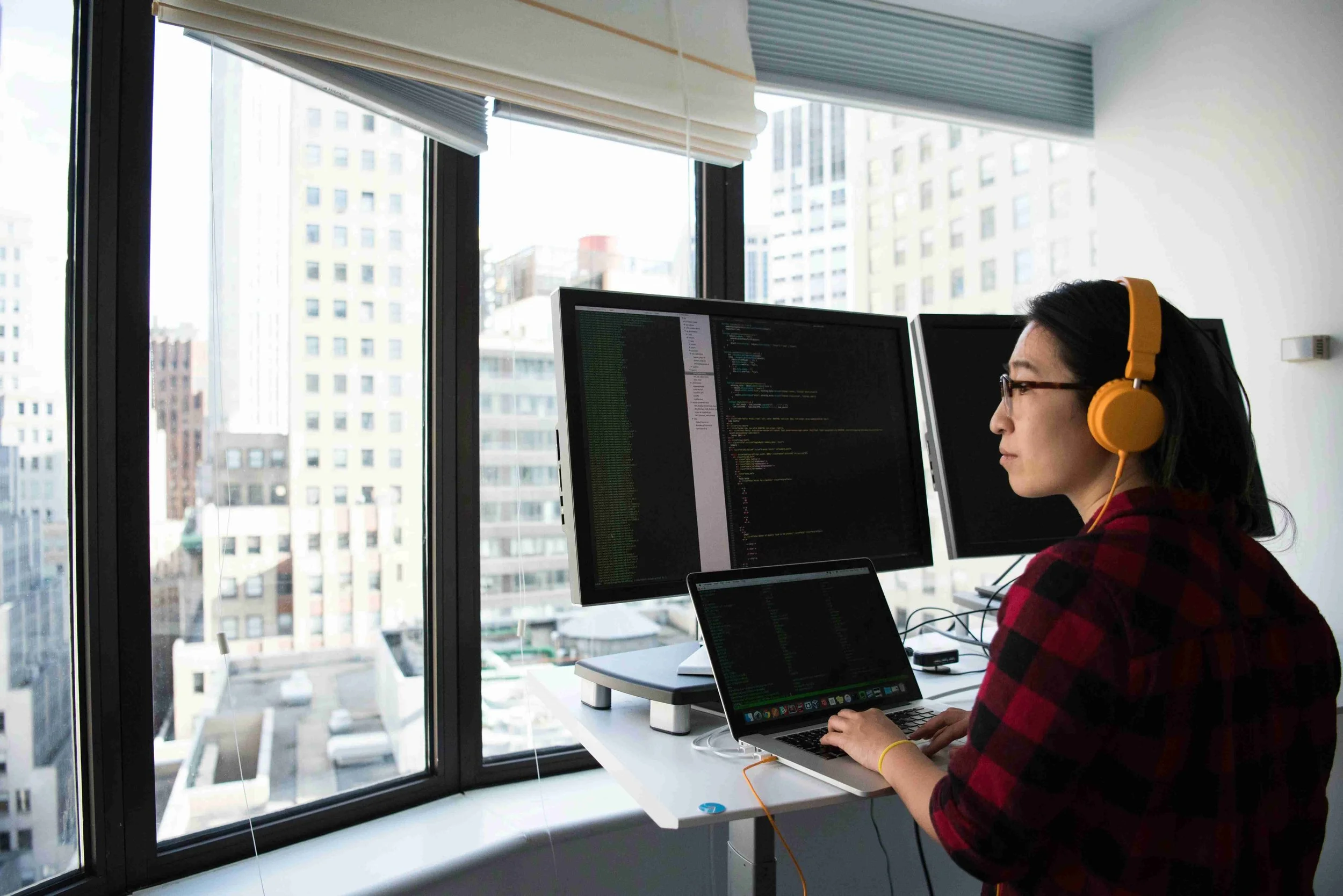 Employee with orange headphones working alone on laptop. They are dressed in a comfortable shirt and are deep in thought looking out the window.