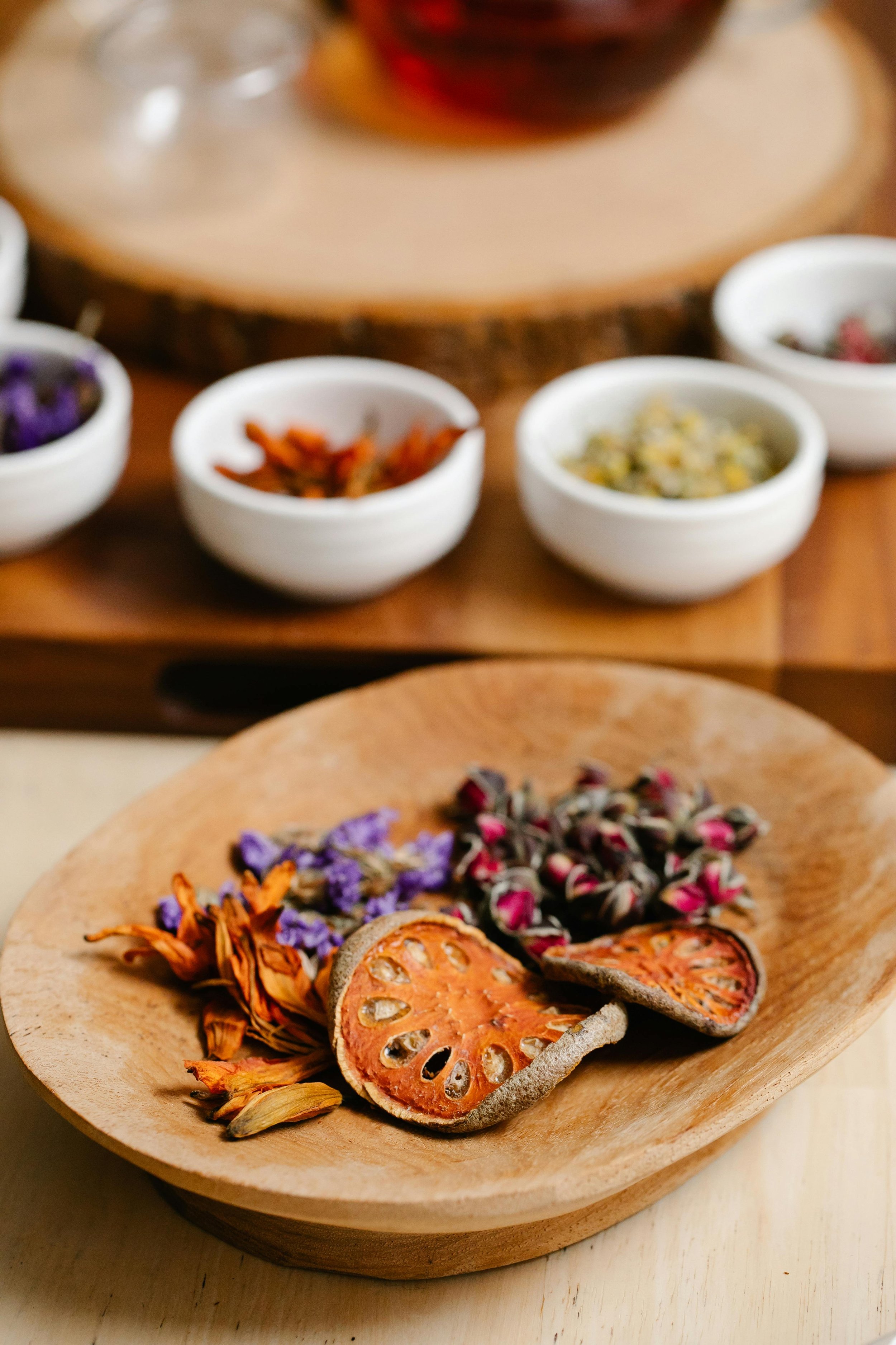 Dried orange slices and assorted dried flowers on a wooden plate with bowls of herbs and spices in the background.