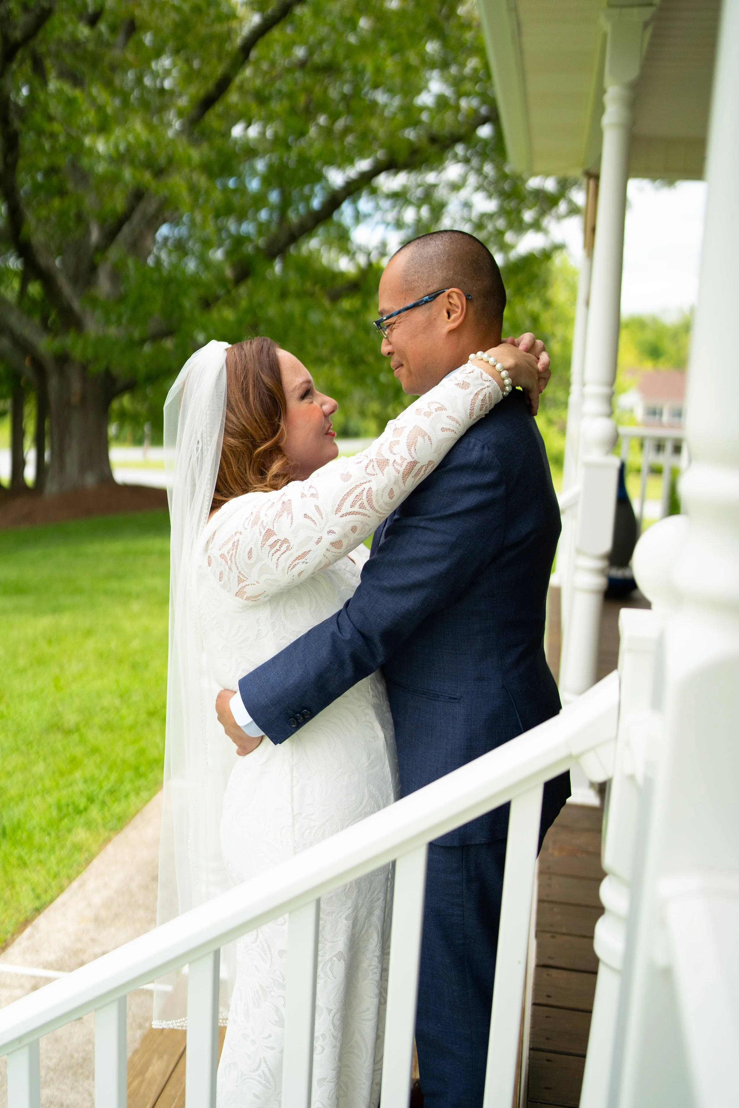 A bride and groom gazing affectionately at each other on a porch with lush green trees in the background.