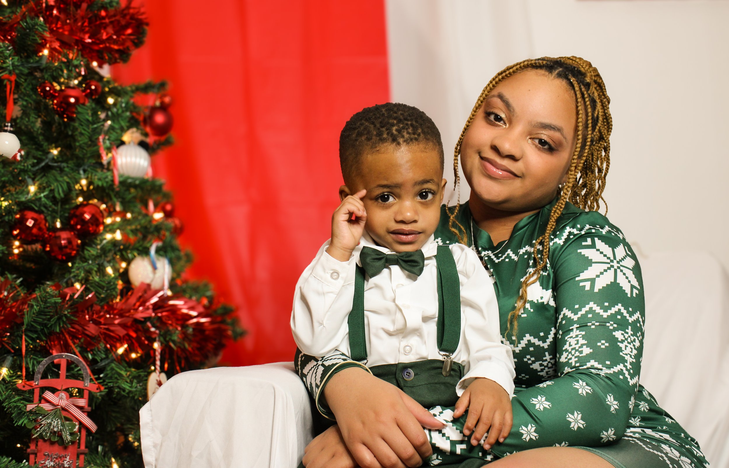 A woman with blond braided hair and a boy with short curly hair sitting on a white couch beside a decorated Christmas tree with red and white ornaments, in front of a red curtain and white background.
