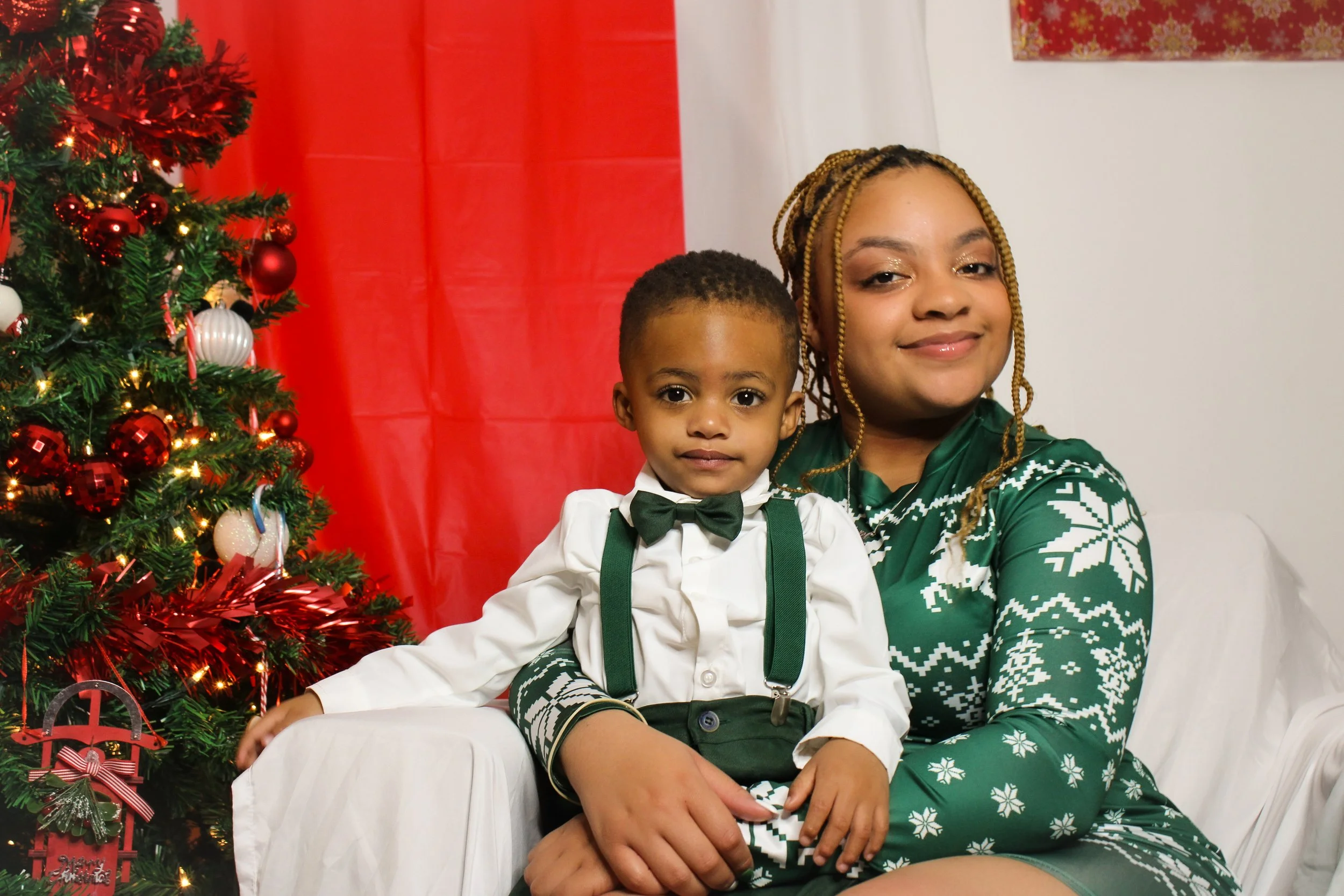A woman and a young boy sitting on a white sofa next to a decorated Christmas tree, smiling at the camera. The woman is wearing a green holiday sweater with white snowflake patterns, and the boy is dressed in a white shirt with a black bow tie and gr