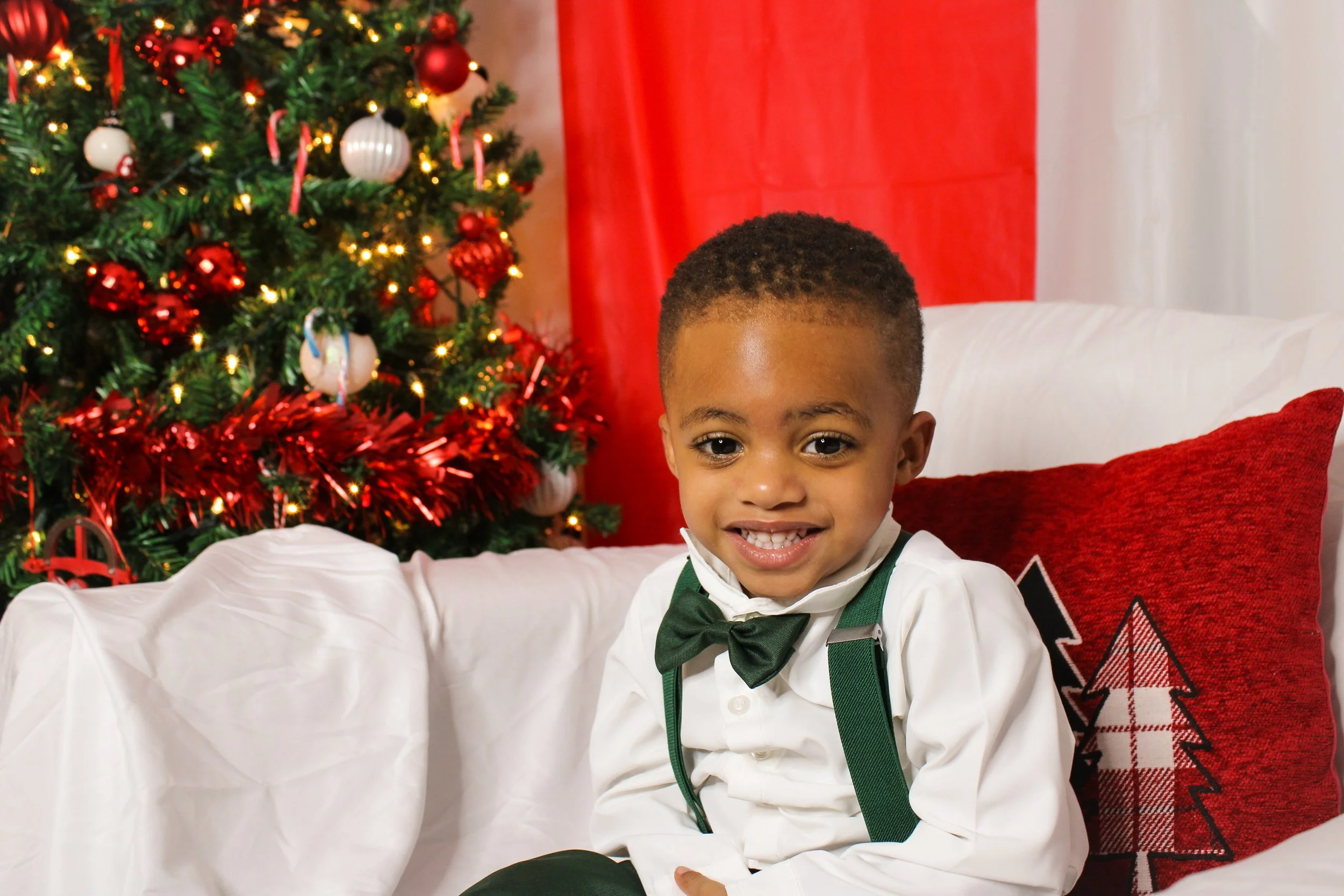A young boy dressed in a white shirt, green suspenders, and a green bow tie sitting on a white sofa, smiling, with a decorated Christmas tree in the background and a red pillow with a Christmas tree design.