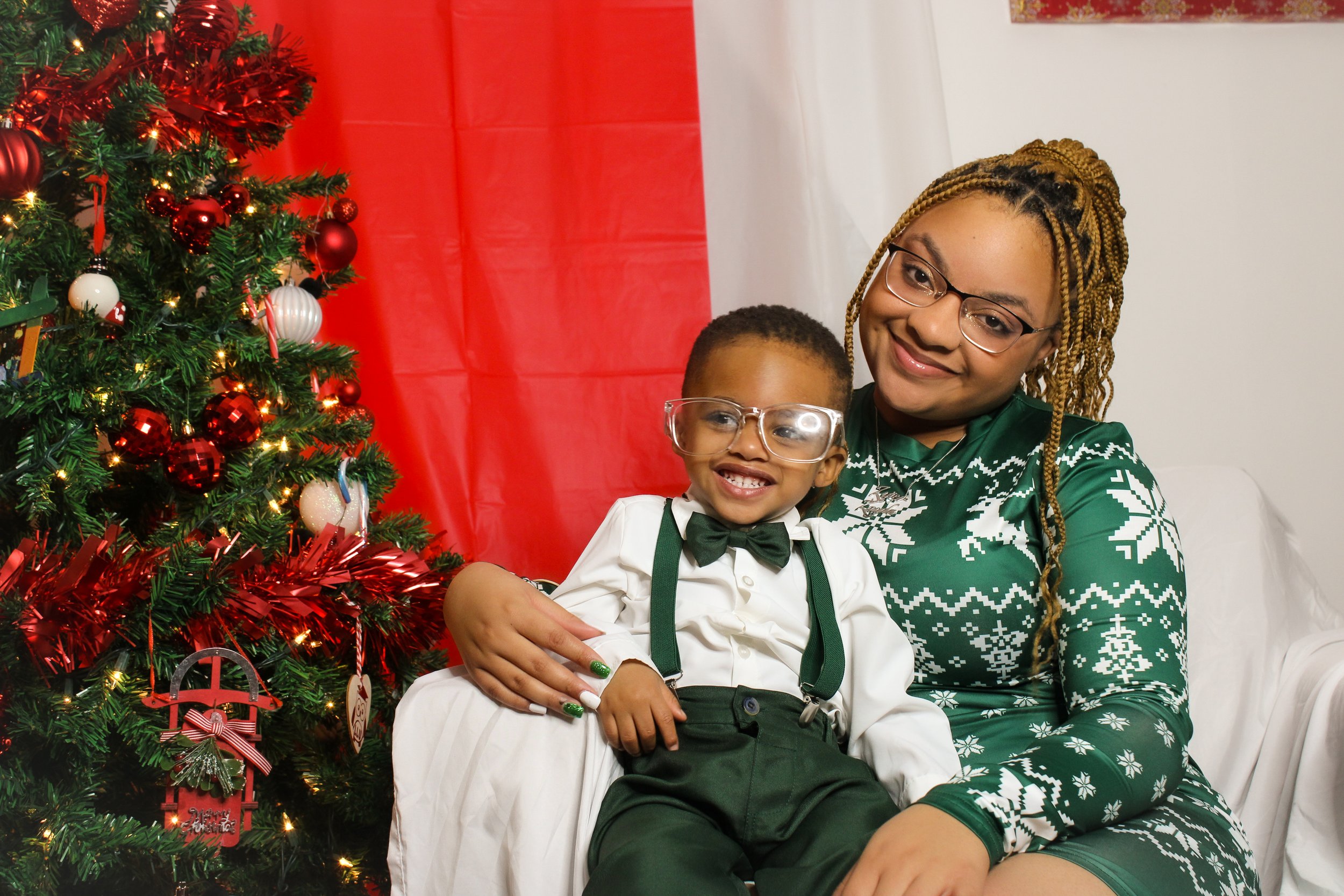 A woman and a young boy sitting together next to a decorated Christmas tree, smiling. The woman is wearing a green Christmas sweater with white snowflake patterns, and the boy is dressed in a white shirt, green suspenders, and a black bow tie.