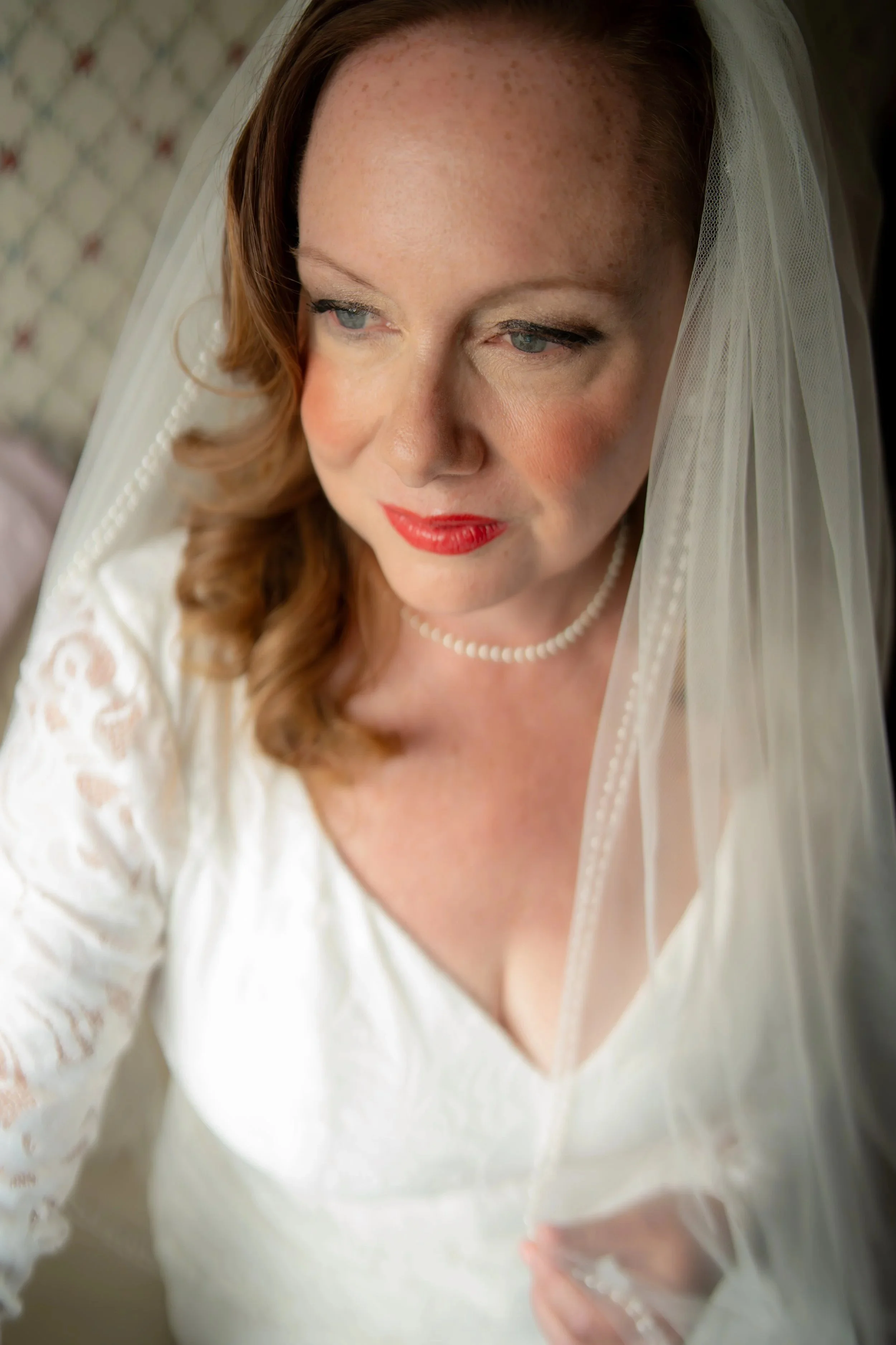 Close-up of a bride with red hair, wearing a white lace wedding dress, a pearl necklace, and a veil, looking down thoughtfully.
