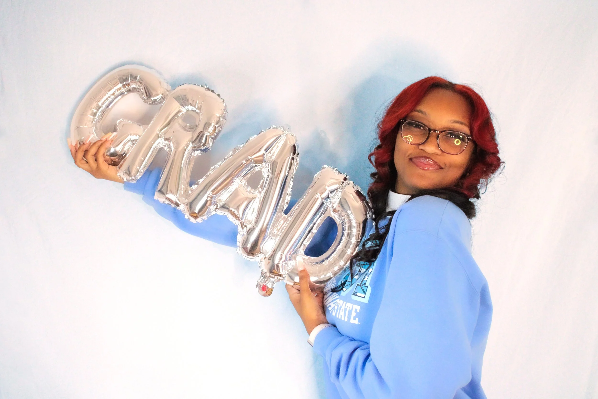 A woman with red hair and glasses holding silver balloons spelling 'GRAD' against a light background.