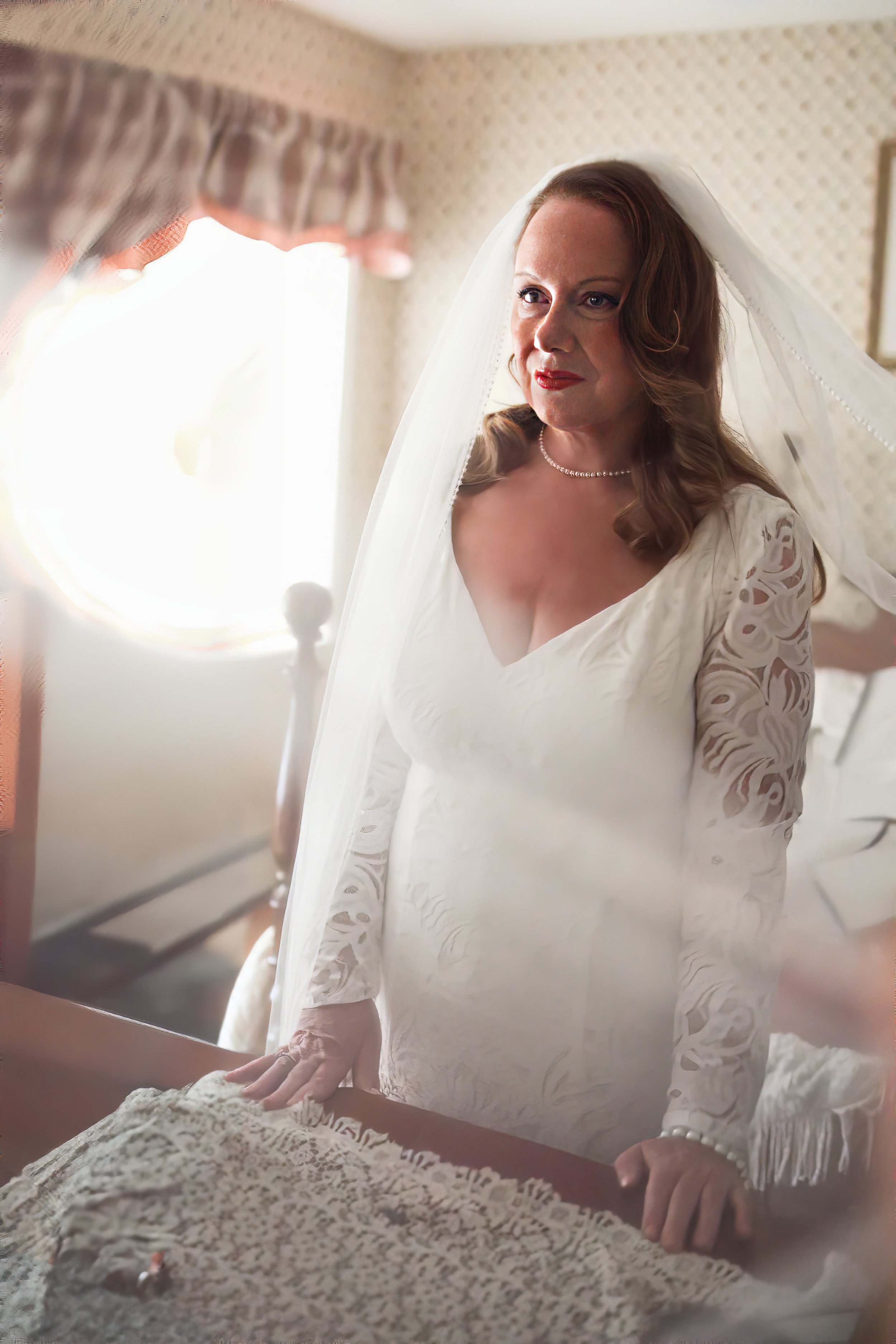 A woman dressed as a bride, wearing a white dress with lace sleeves and a veil, looking at herself in a mirror with sunlight coming through the window behind her.