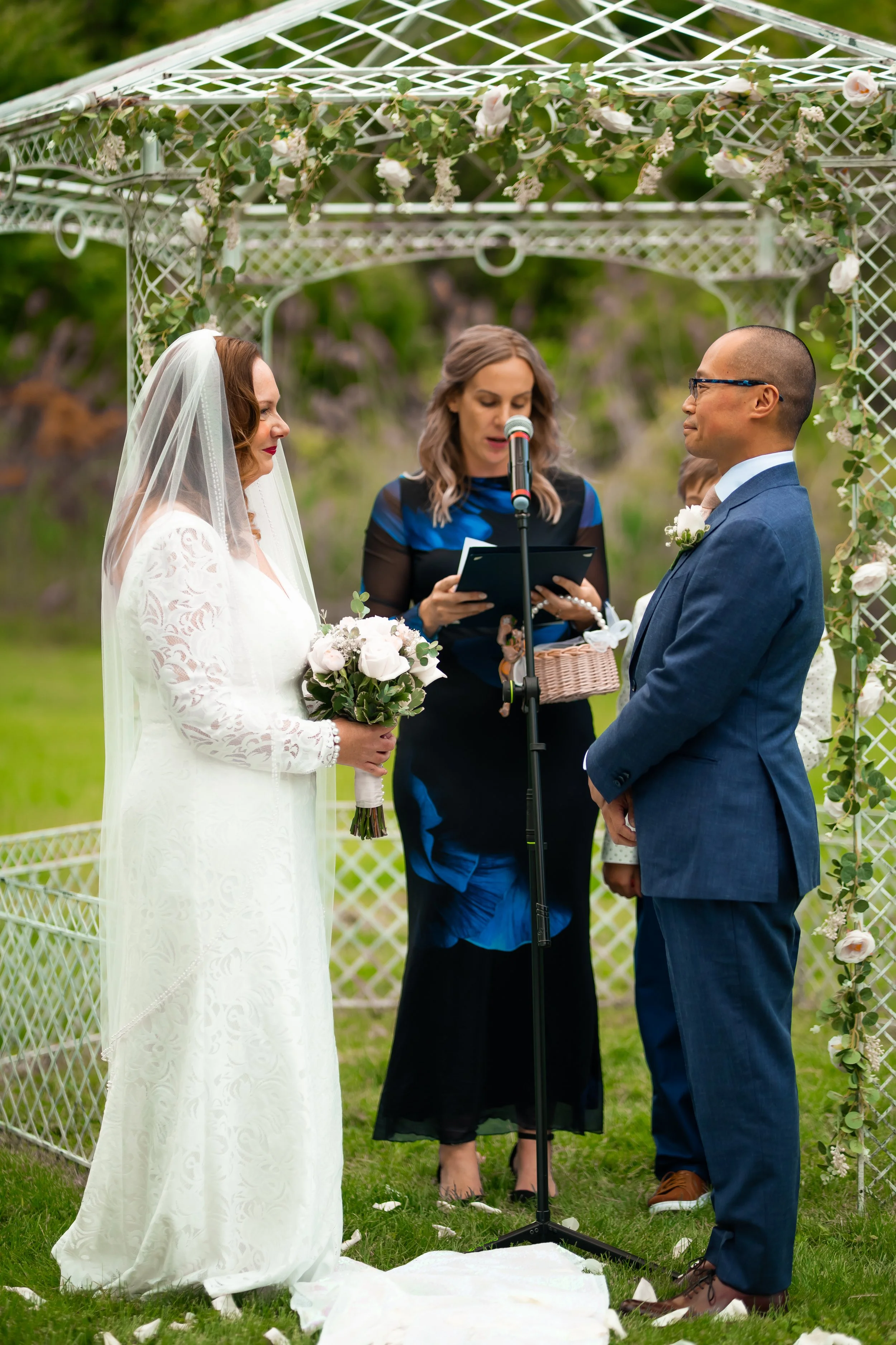 A wedding ceremony with a bride and groom standing under a decorated arch, with an officiant reading vows.