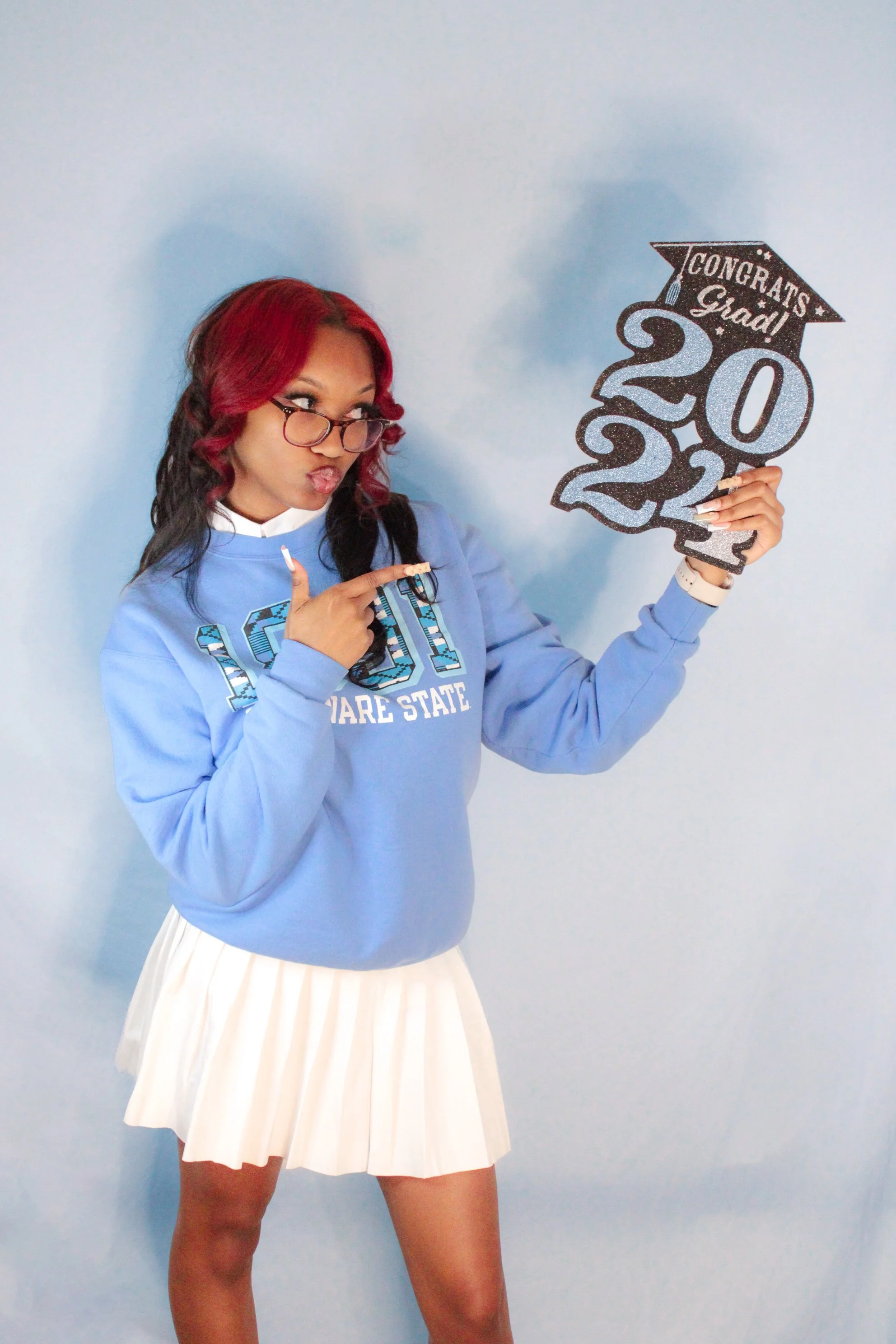 A young woman with red and black hair, glasses, and wearing a blue sweatshirt and white pleated skirt, pointing at a graduation sign with the year 2022, indicating her graduation.