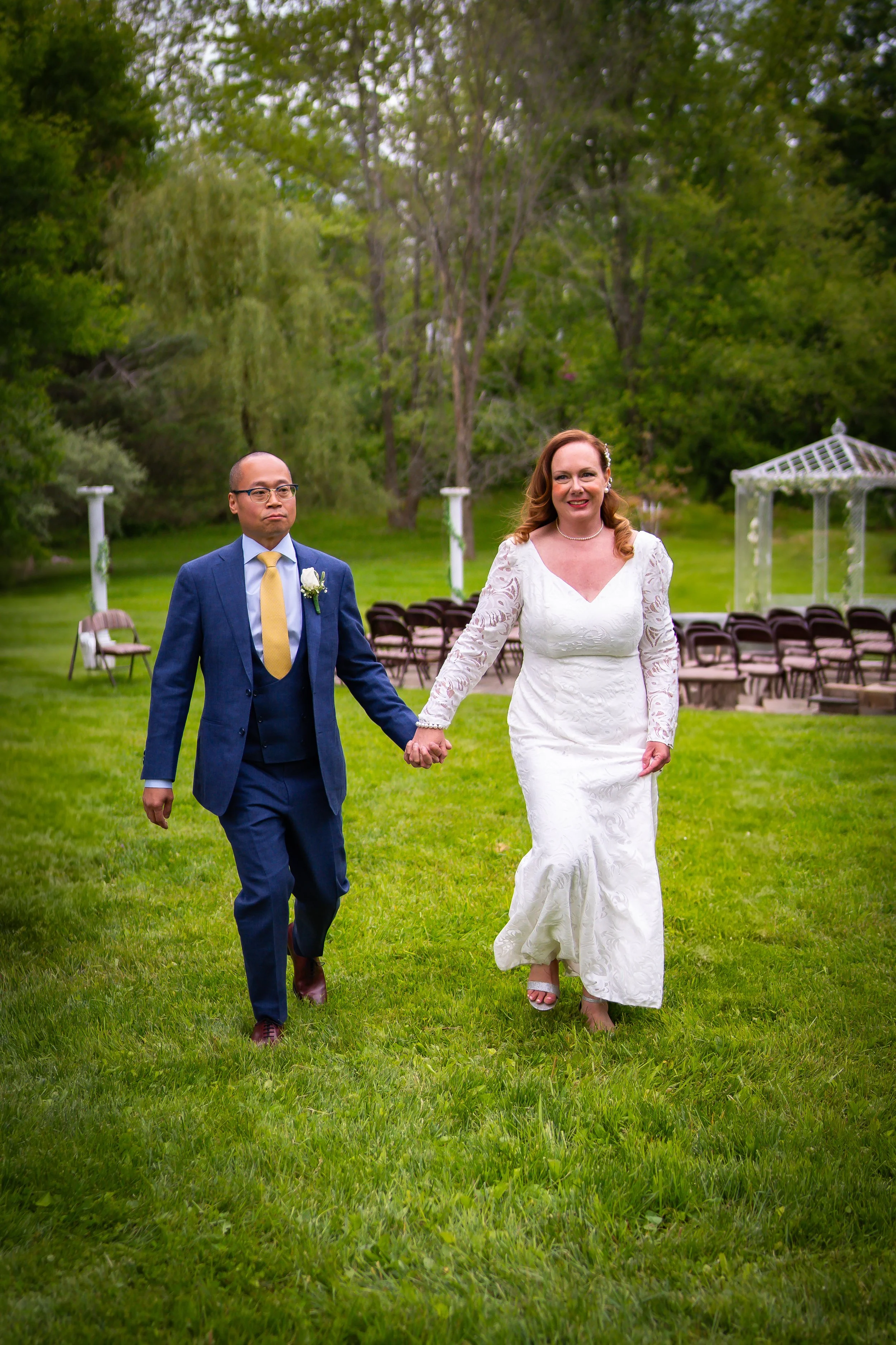 A bride in a white lace wedding gown and a groom in a blue suit walking hand in hand outdoors on a grassy area.