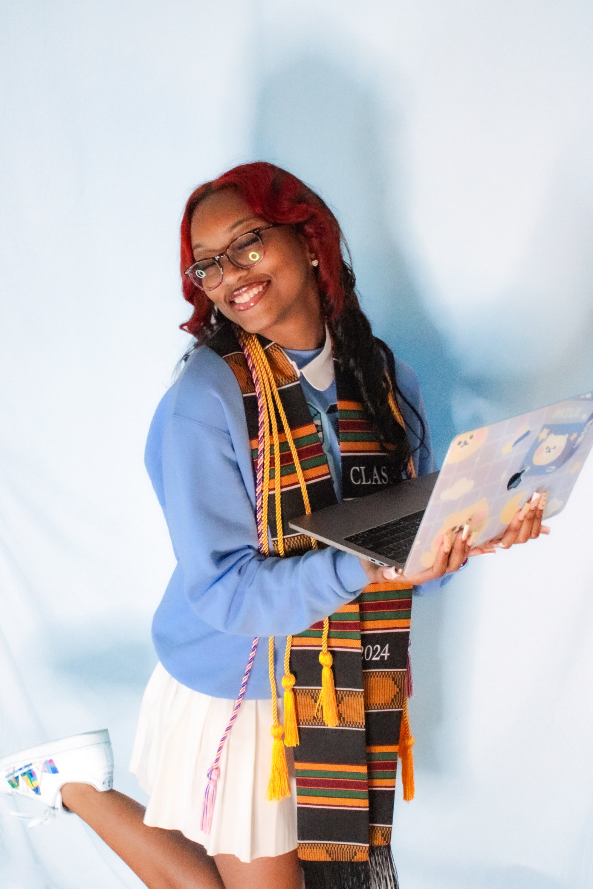 A young woman in a graduation gown and colorful stole, holding a laptop, smiling against a light blue background.
