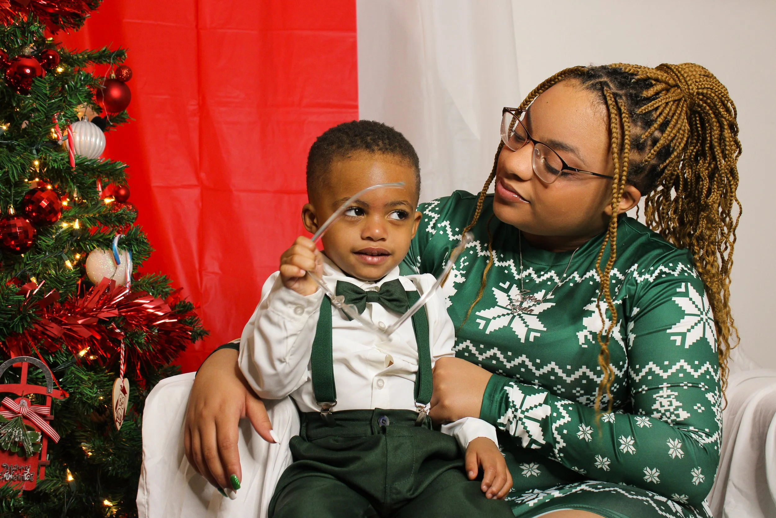 A woman and a young boy sitting by a decorated Christmas tree, with the woman looking at the boy who is holding a Christmas ornament.