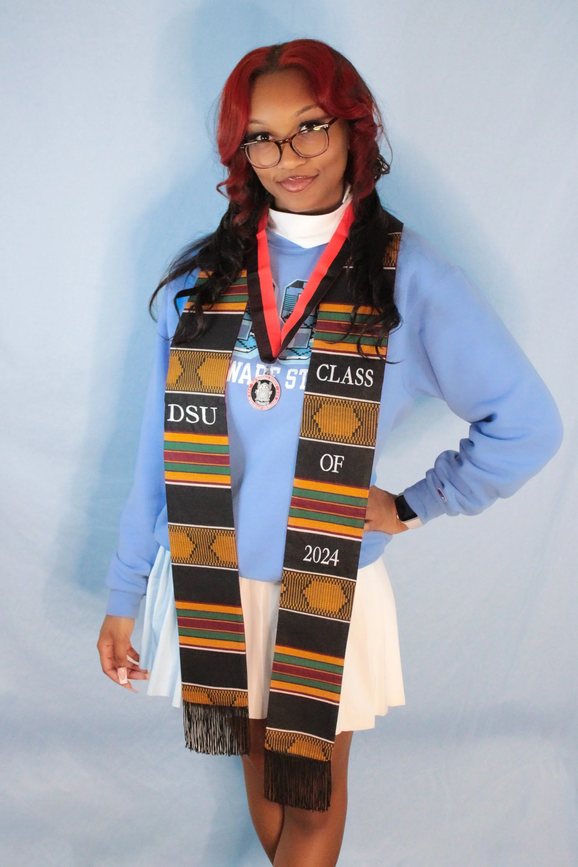Young woman with red and black hair, glasses, wearing a blue sweatshirt, and draped with a black and orange patterned graduation stole. She has a medal around her neck and is standing against a light blue background.