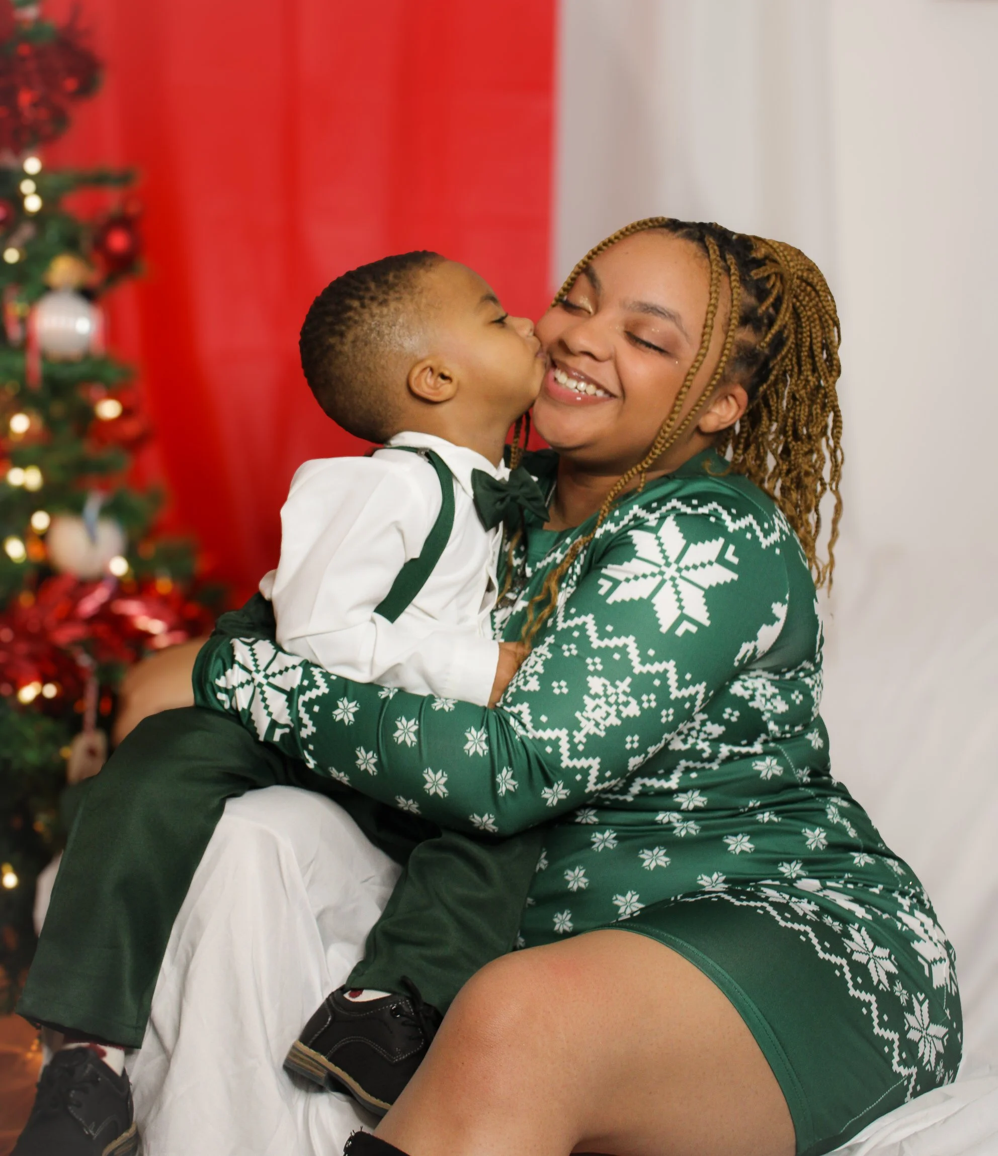 A young boy giving a kiss on the cheek to a woman, who is smiling with her eyes closed, in a festive holiday setting with a decorated Christmas tree in the background.