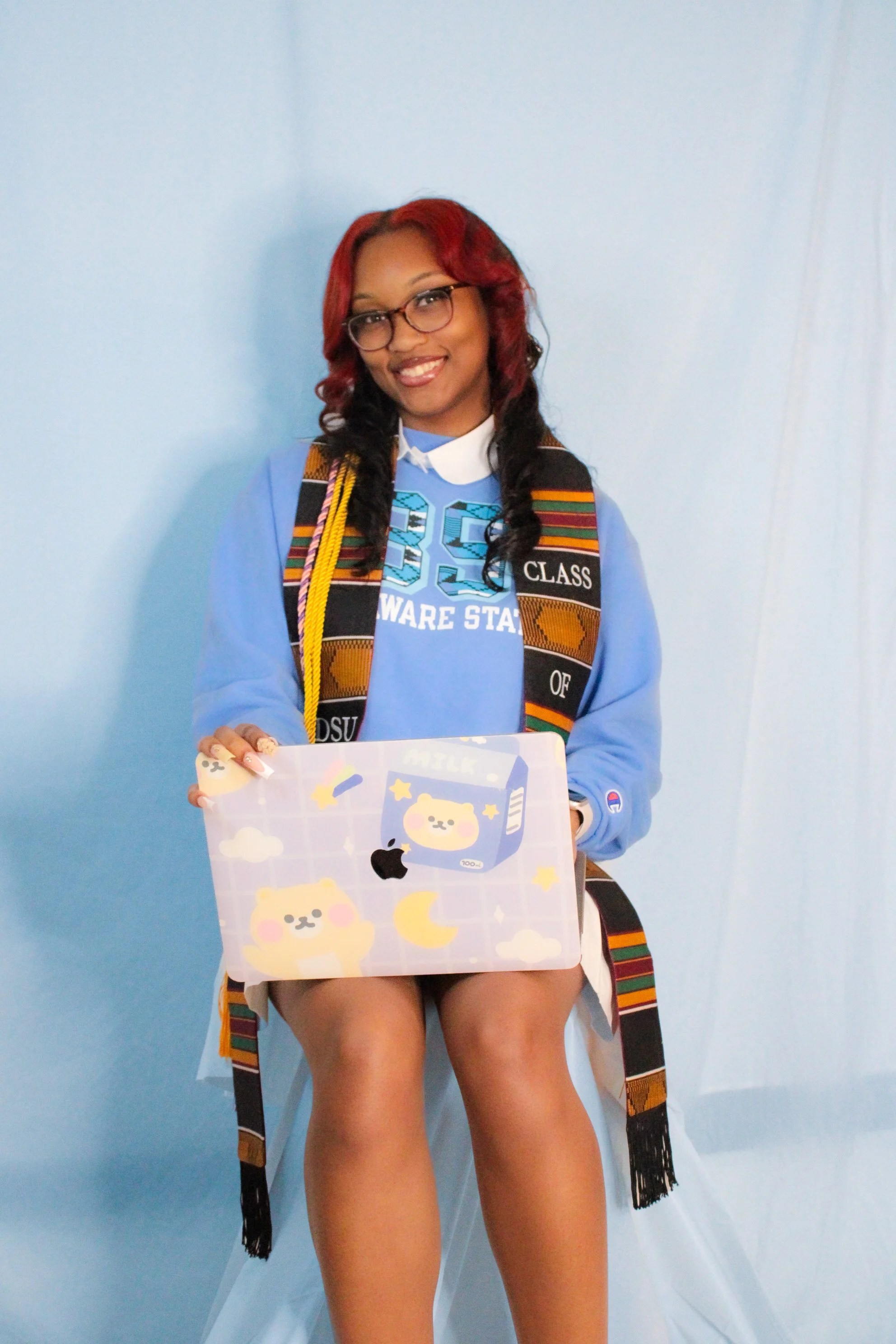 A young woman with red and black curly hair wearing glasses, a blue hoodie with college text, and a decorative sash, sitting on a chair against a light blue background. She holds a laptop covered with cute bear and moon stickers.