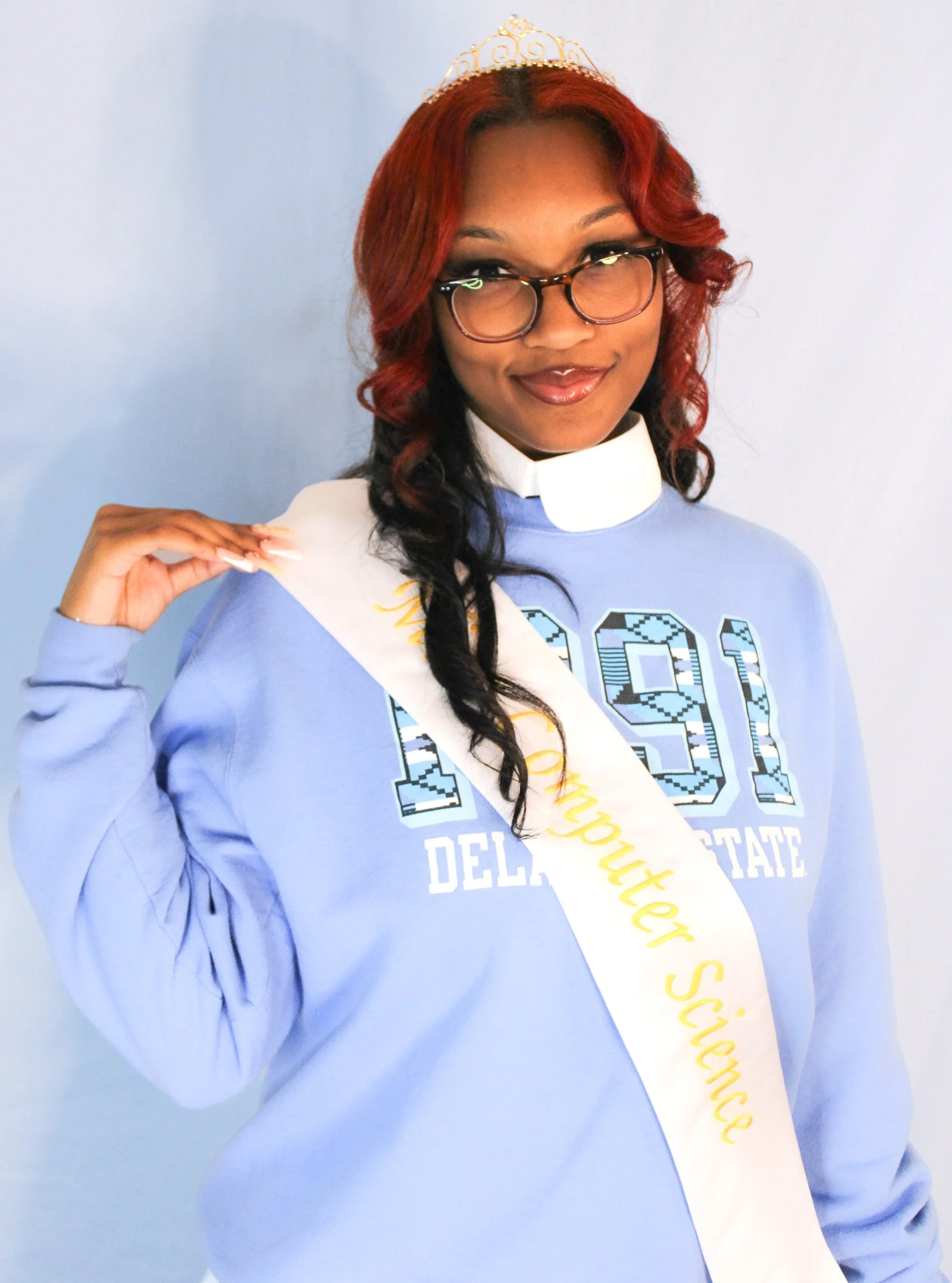 Young woman with curly red and black hair wearing glasses, a blue sweatshirt with '1971 Delaware State' printed on it, a white sash that reads 'Missour Science', and a gold tiara, posing against a light blue background.