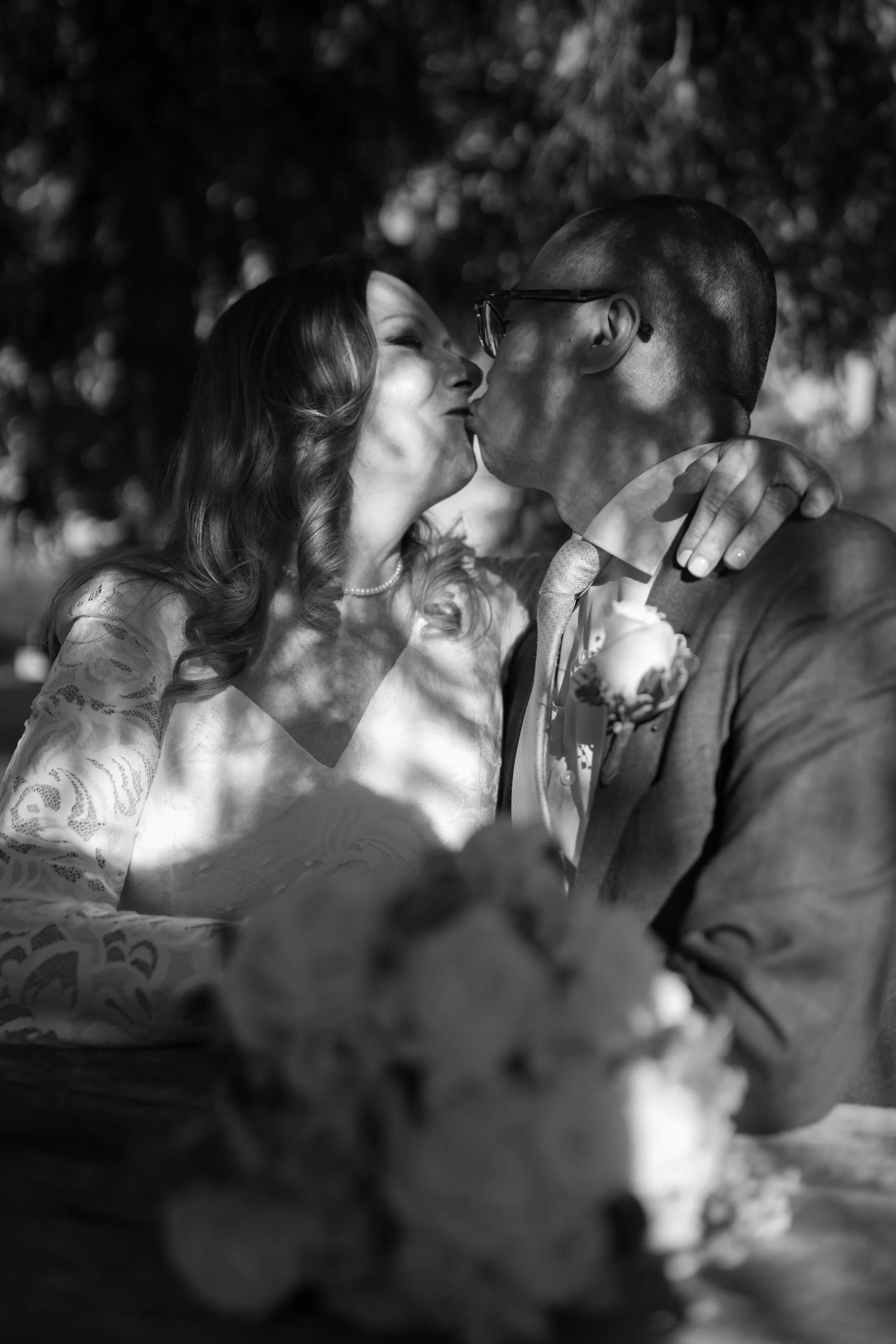 Black and white photo of a bride and groom sharing a kiss outdoors, with a blurred wedding bouquet in the foreground.