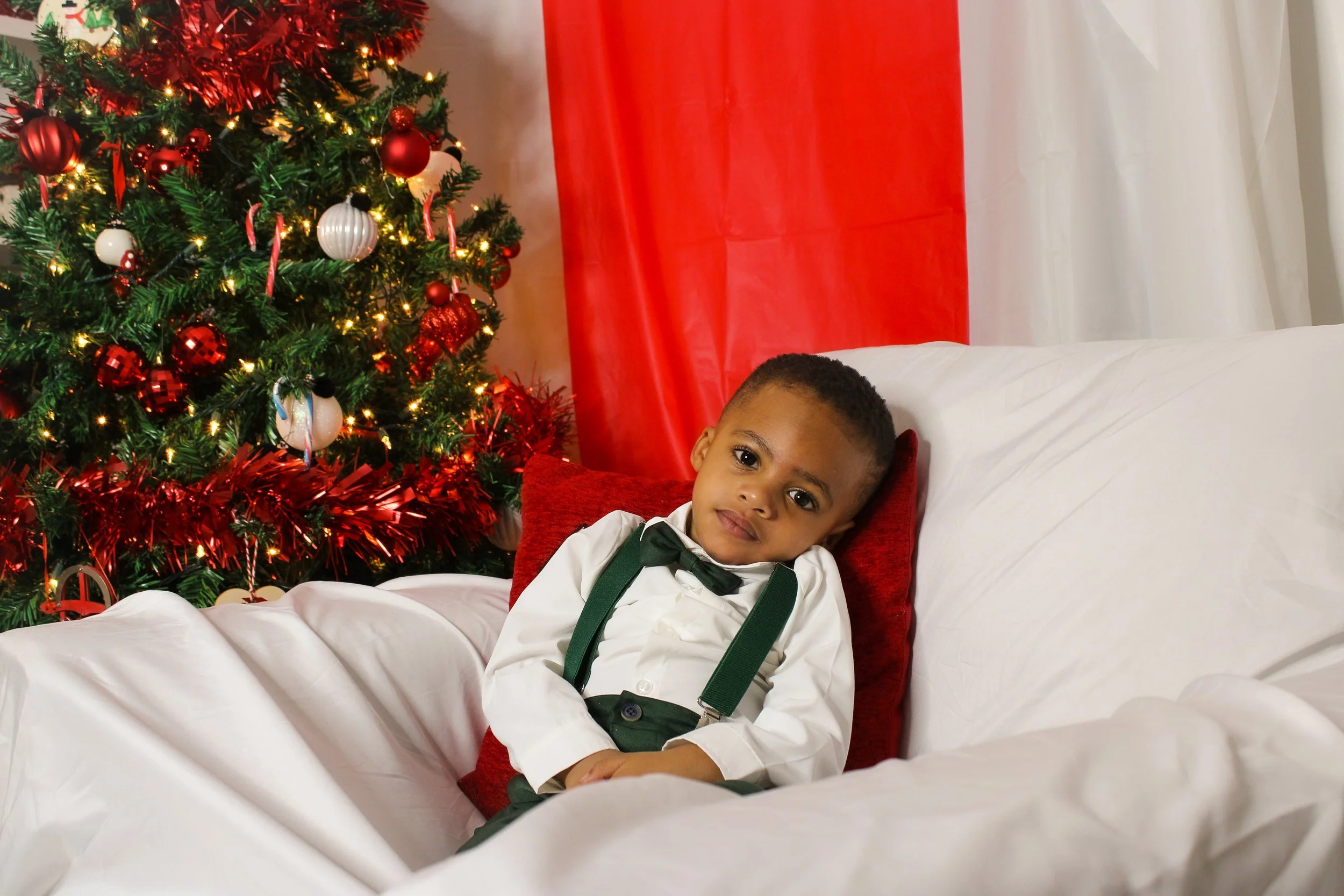 A young boy with curly hair dressed in a white shirt with a black bow tie and suspenders sitting on a sofa, leaning against a red pillow, beside a decorated Christmas tree with ornaments and lights.