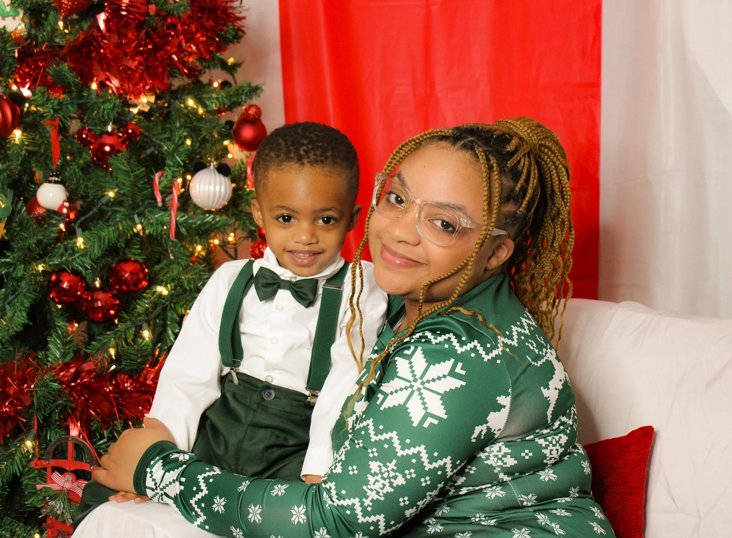 A woman with braids, glasses, and a green Christmas sweater, sitting on a white sofa with a young boy in a white shirt, green suspenders, and a bow tie, in front of a decorated Christmas tree.