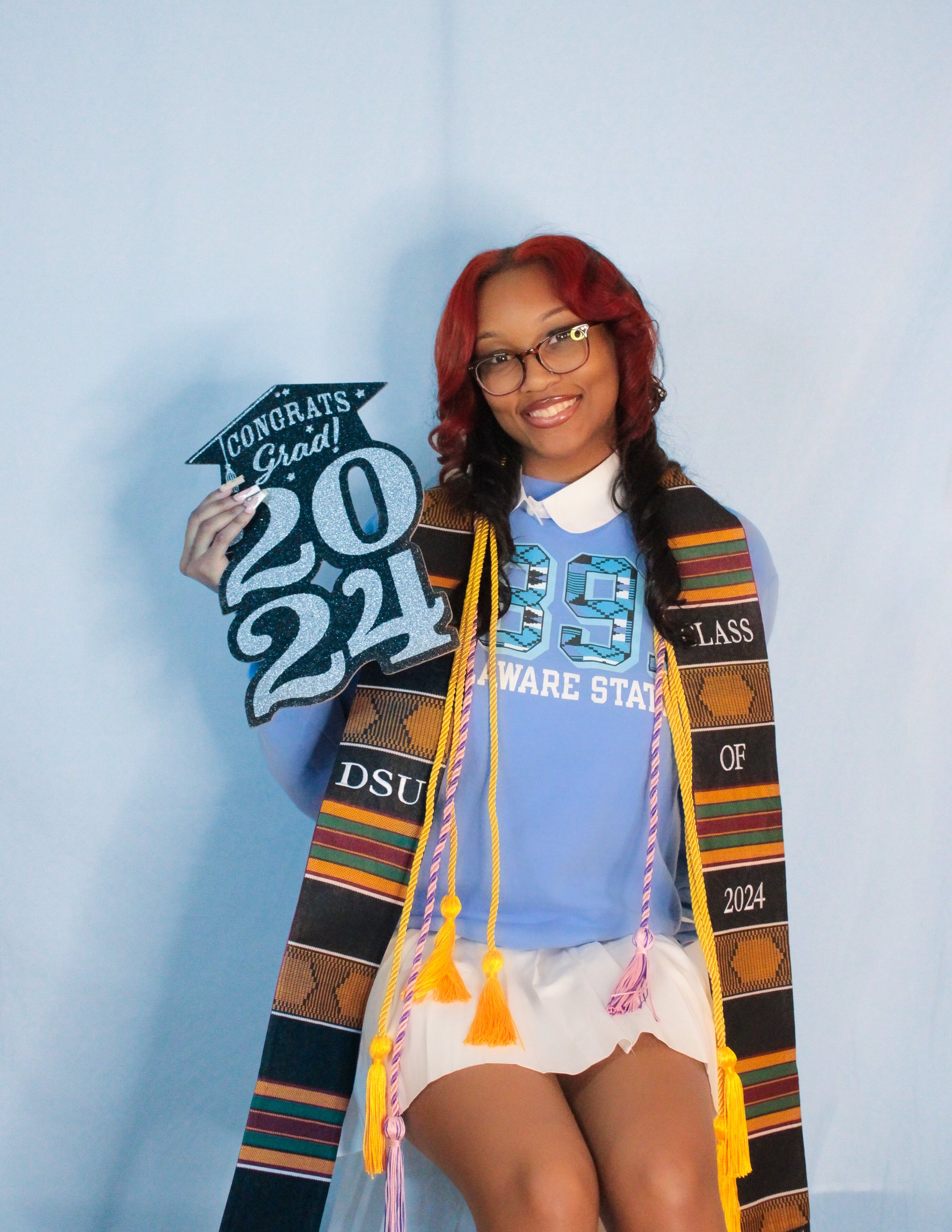 Graduation woman in a blue shirt holding a 'Congrats Grad 2024' sign, wearing a graduation stole and cords, smiling, sitting in front of a plain wall.