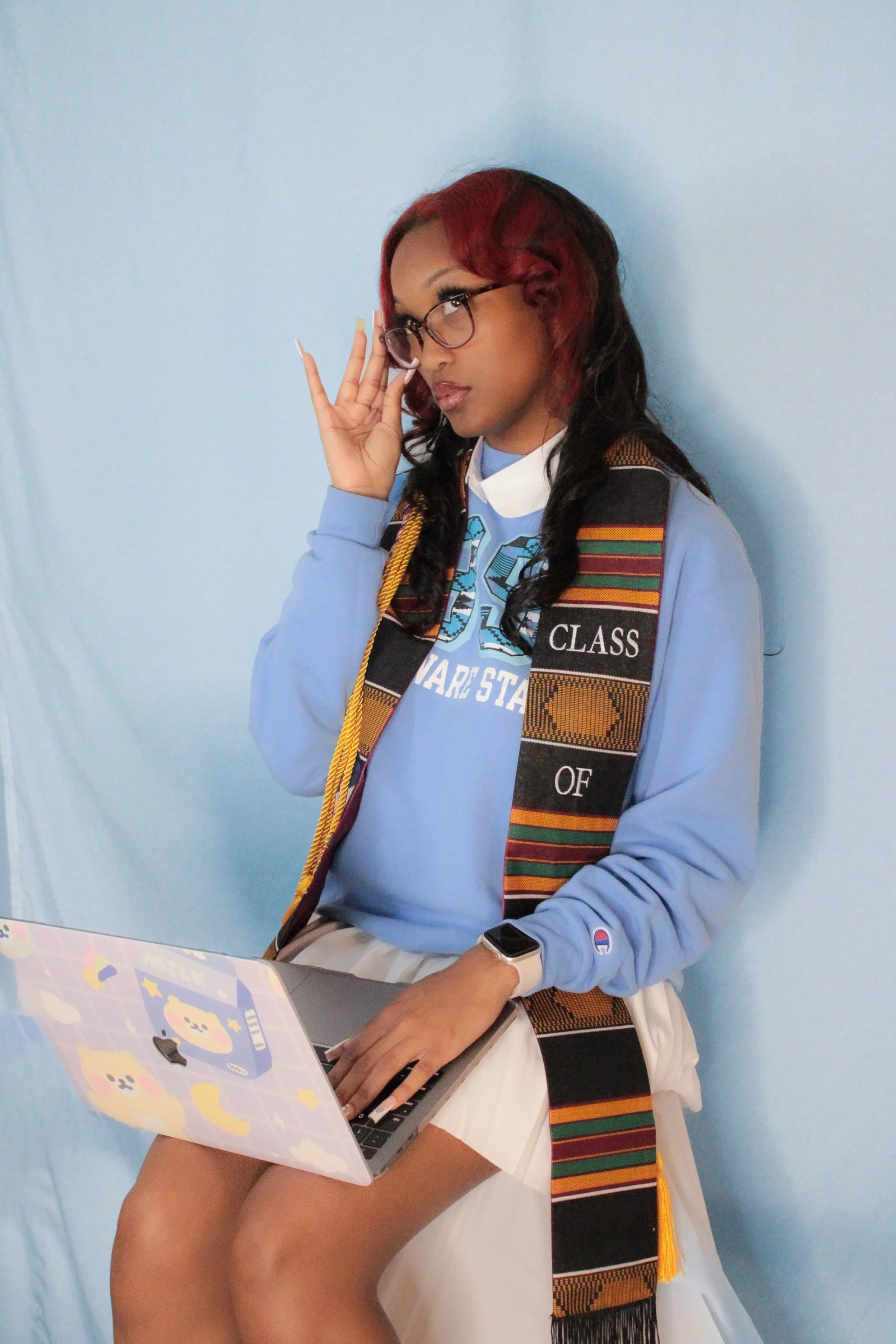 A young woman with red and black hair wearing glasses, a blue hoodie, and a graduation sash, sitting against a light blue wall, holding a laptop decorated with cute character stickers.