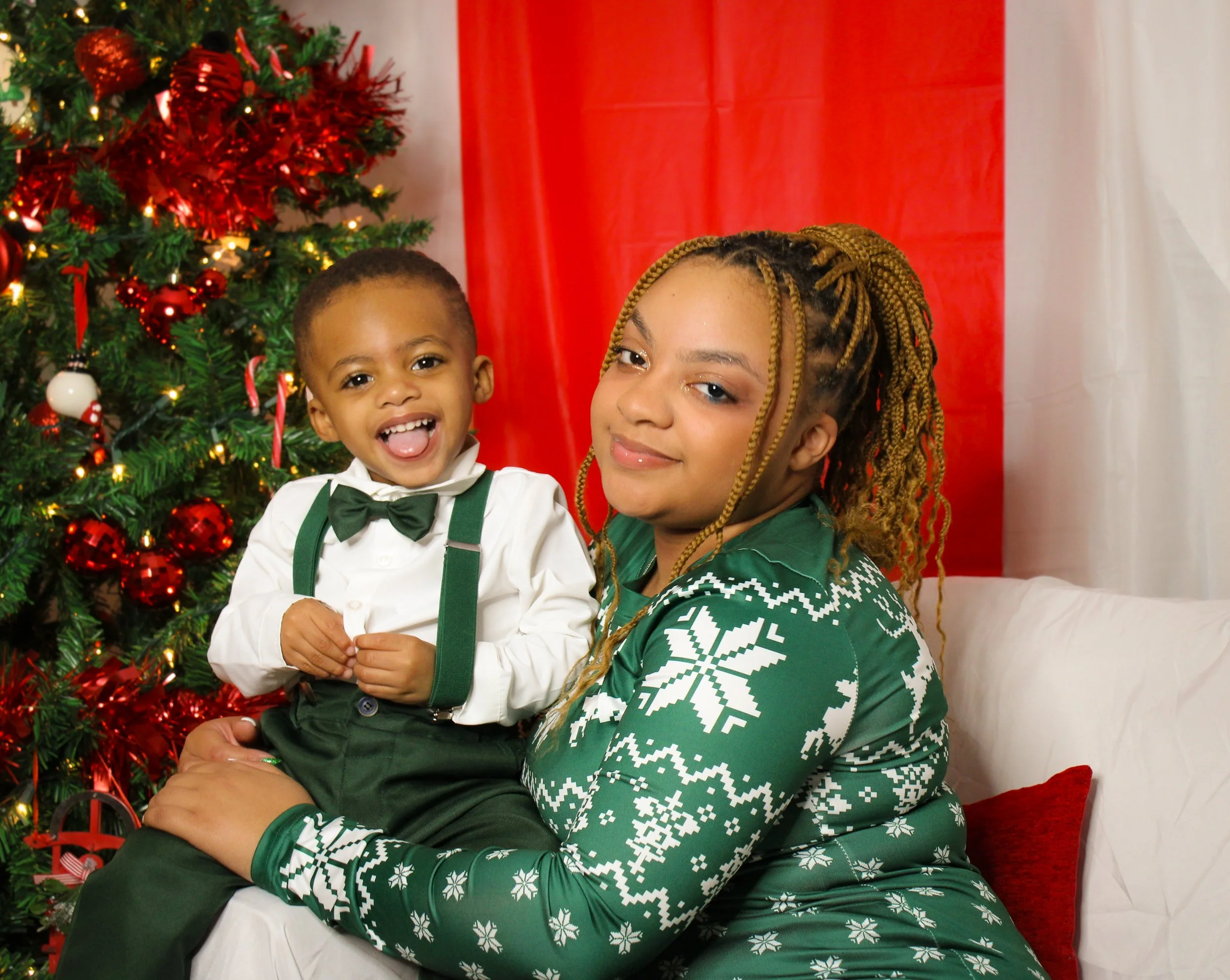 A woman and a young boy celebrating Christmas next to a decorated Christmas tree with red and gold ornaments and lights. The woman wears a green Christmas sweater with white snowflake patterns, and the boy wears a white shirt with a black bow tie, gr