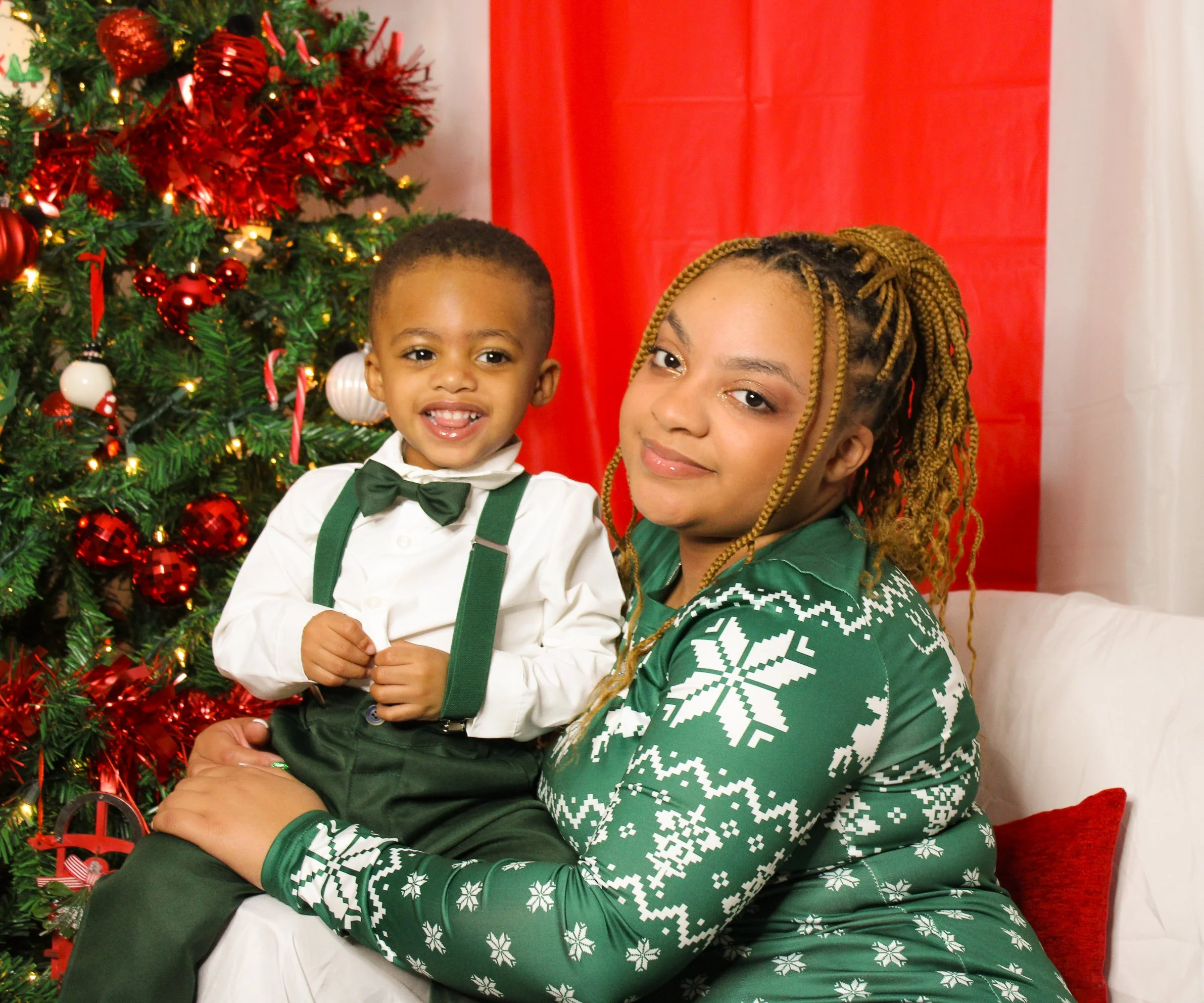 A young boy in a white shirt, black pants with suspenders, and a bow tie, sitting on a woman's lap, smiling in front of a decorated Christmas tree with red and white ornaments. The woman, with braided hair, is wearing a green Christmas sweater with w