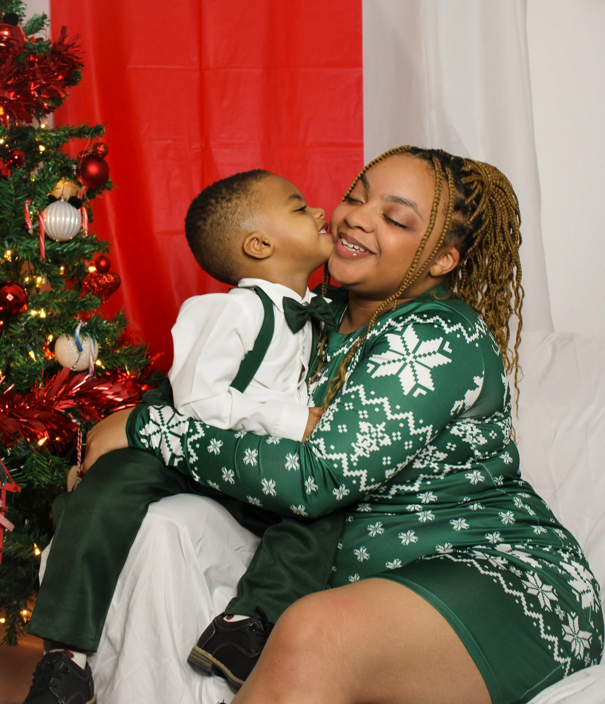A young boy in a tuxedo with a bow tie and suspenders kisses a smiling woman in a green holiday sweater with snowflake patterns, sitting near a decorated Christmas tree.