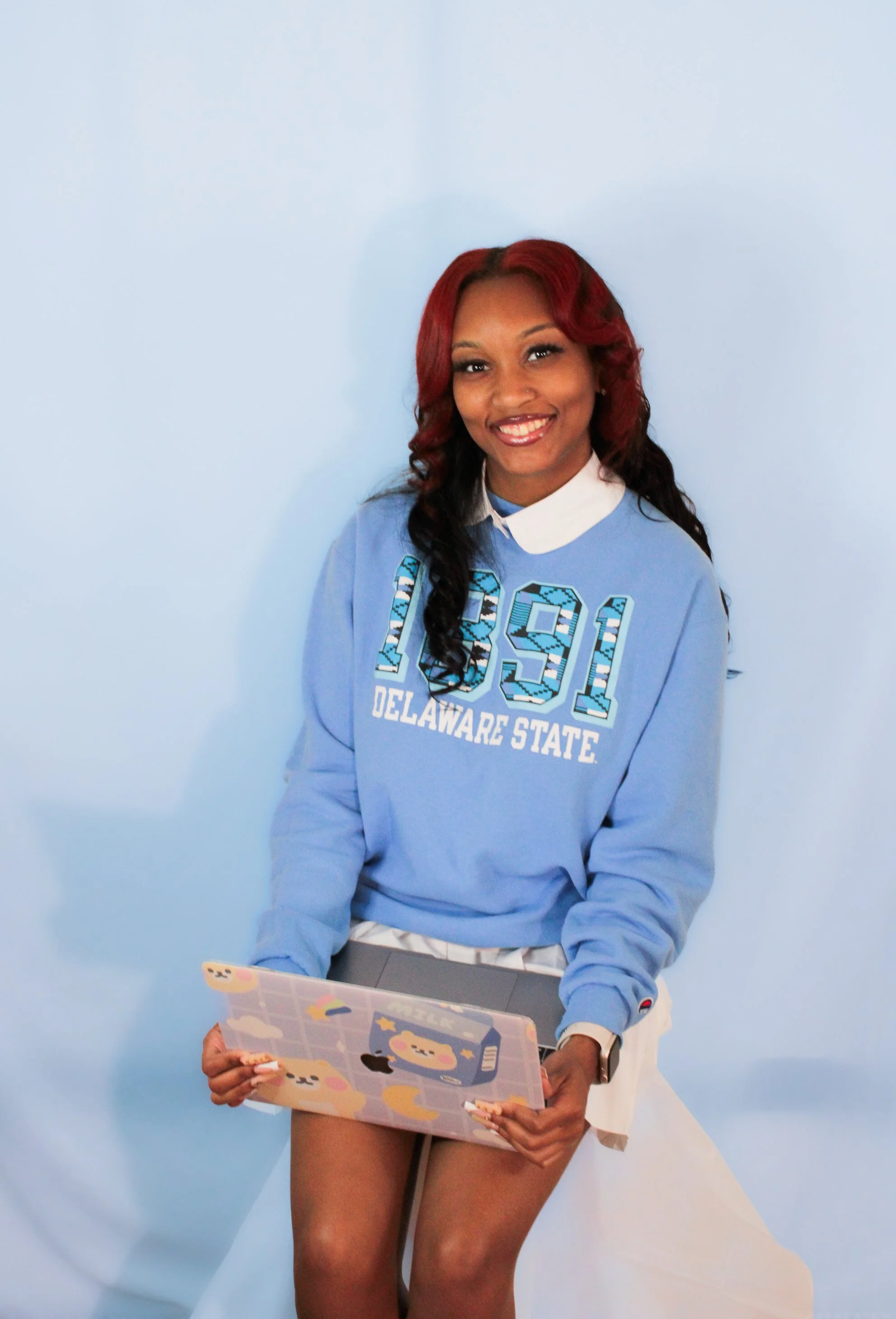 A young woman with red hair sitting against a light blue background, wearing a blue Delaware State sweatshirt and holding a laptop decorated with bear stickers.