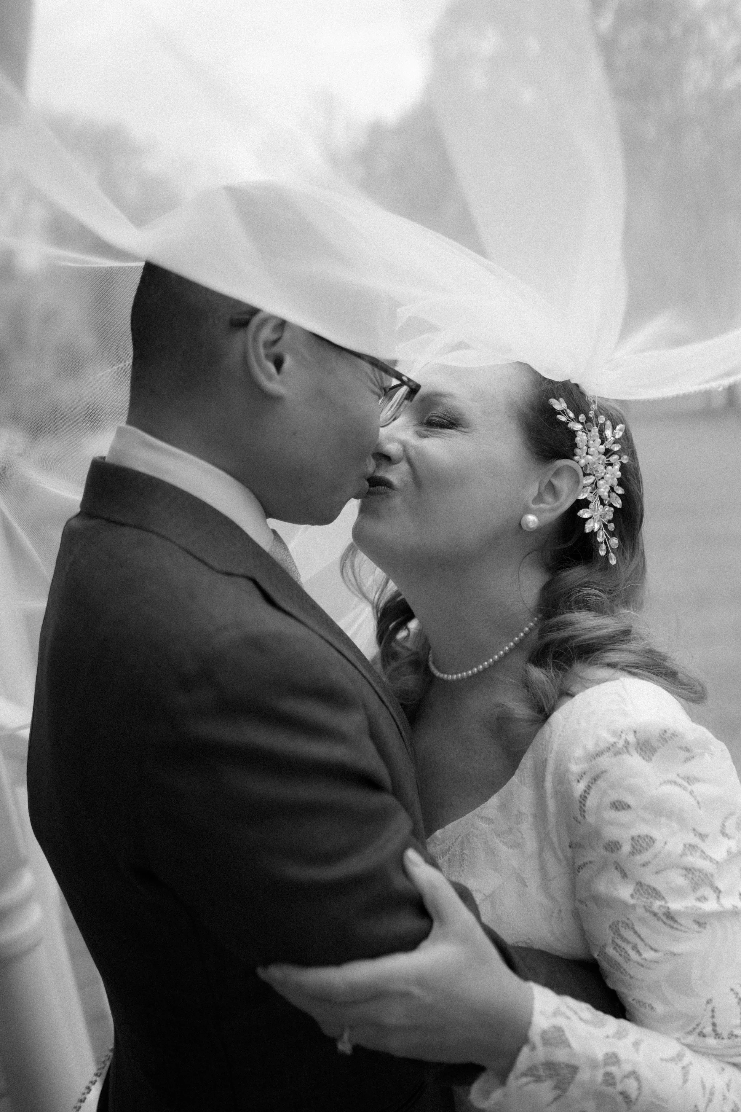 Black and white photo of a newlywed couple sharing a kiss, with the bride wearing a lacy dress, pearl earrings, a pearl necklace, and an ornate headpiece, while the groom is in a suit wearing glasses, under a large hat or veil.