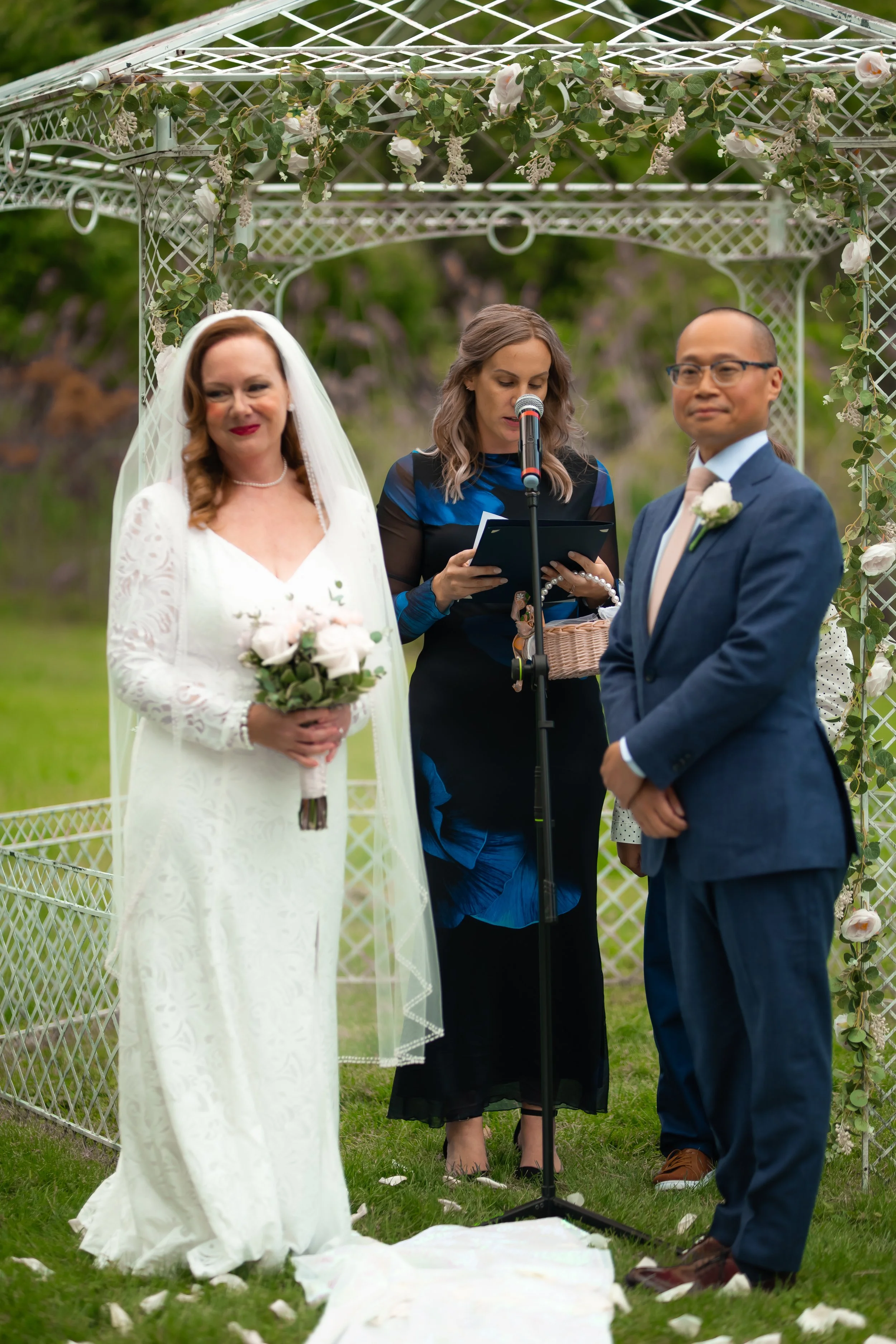 A wedding ceremony outdoors with a bride in a white lace gown and veil holding a bouquet, a groom in a blue suit, and a woman officiant reading from a book under a decorated arch with flowers.