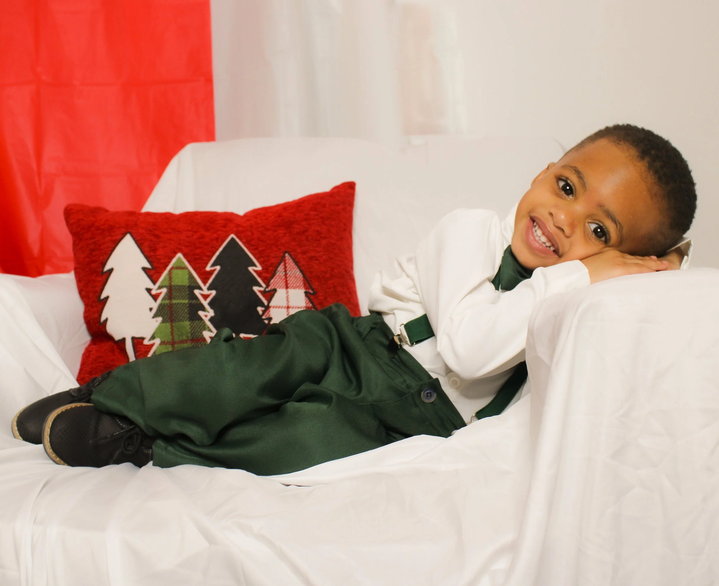 A smiling young boy lying on a white couch, resting his head on his arms, with a red pillow featuring Christmas tree patterns behind him, and red and white curtains in the background.