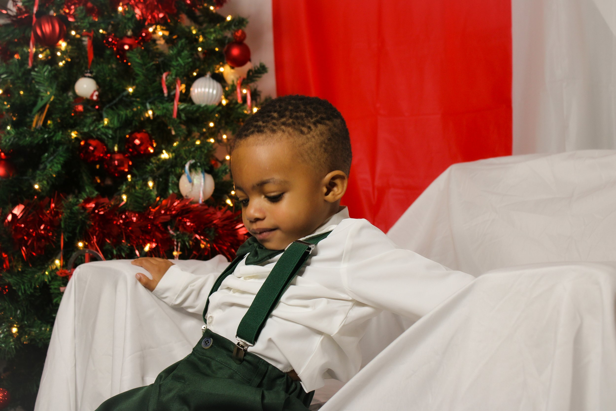 A young boy dressed in white shirt with green suspenders sitting on a white chair in front of a decorated Christmas tree with ornaments and lights.
