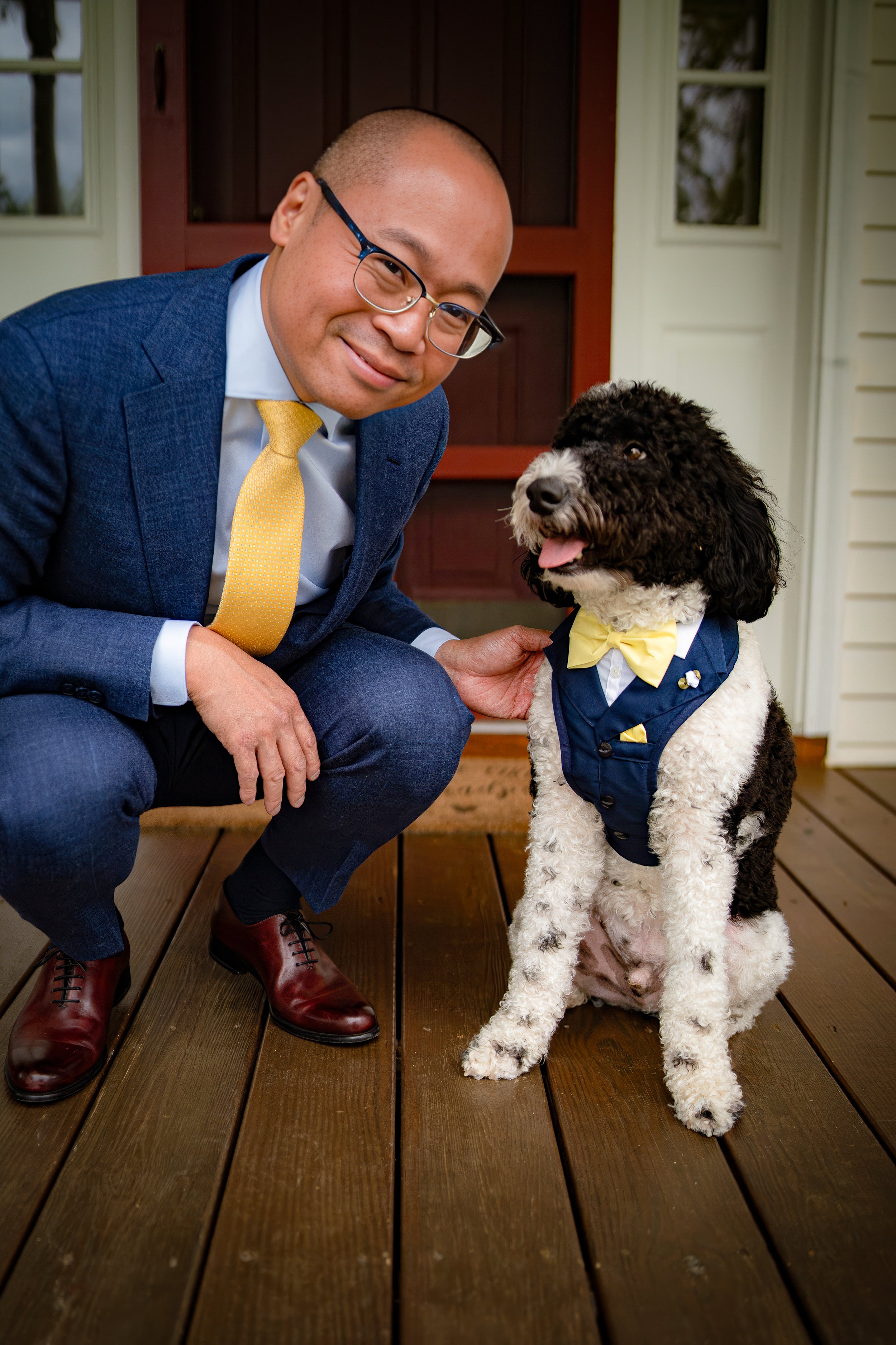 Man in suit and glasses kneeling next to a black and white dog wearing a blue tuxedo with yellow bow tie on a wooden porch.