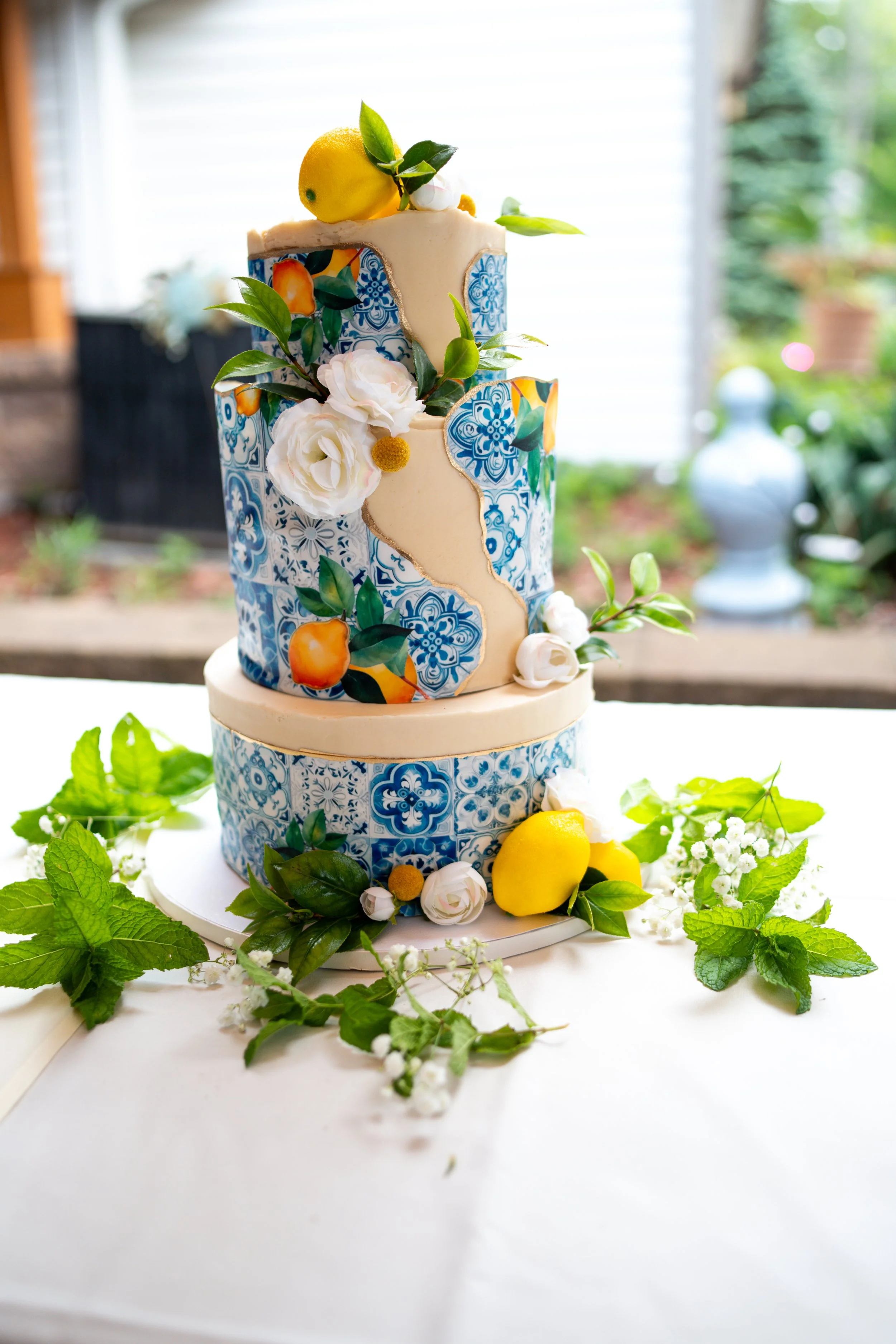 Two-tiered decorative cake with blue and white mosaic tile pattern, adorned with white flowers, green leaves, and bright yellow lemons, set on a white table with greenery surrounding it.
