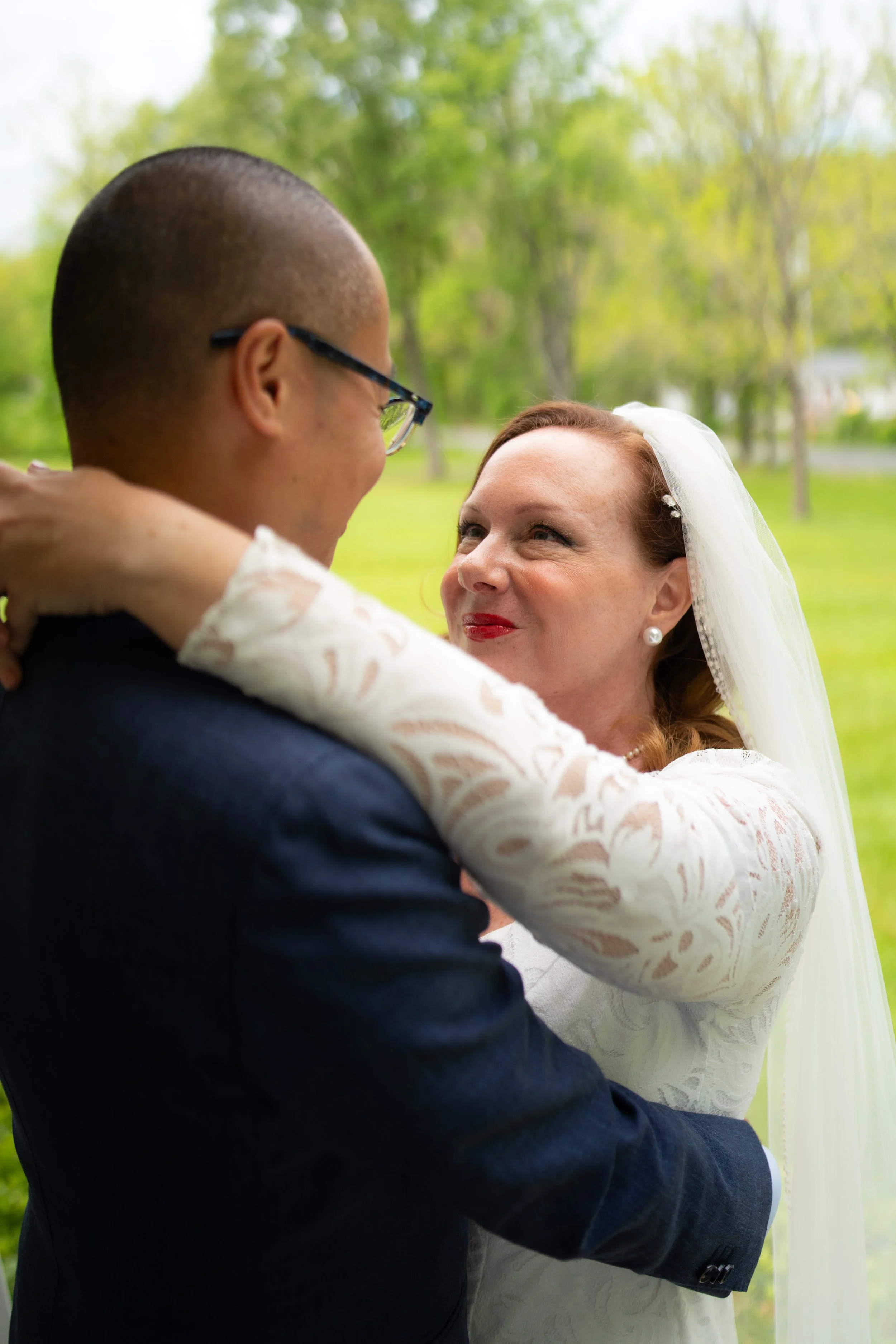 A bride with red hair and a white veil embracing a groom with glasses and a dark suit outdoors in a green, leafy area.