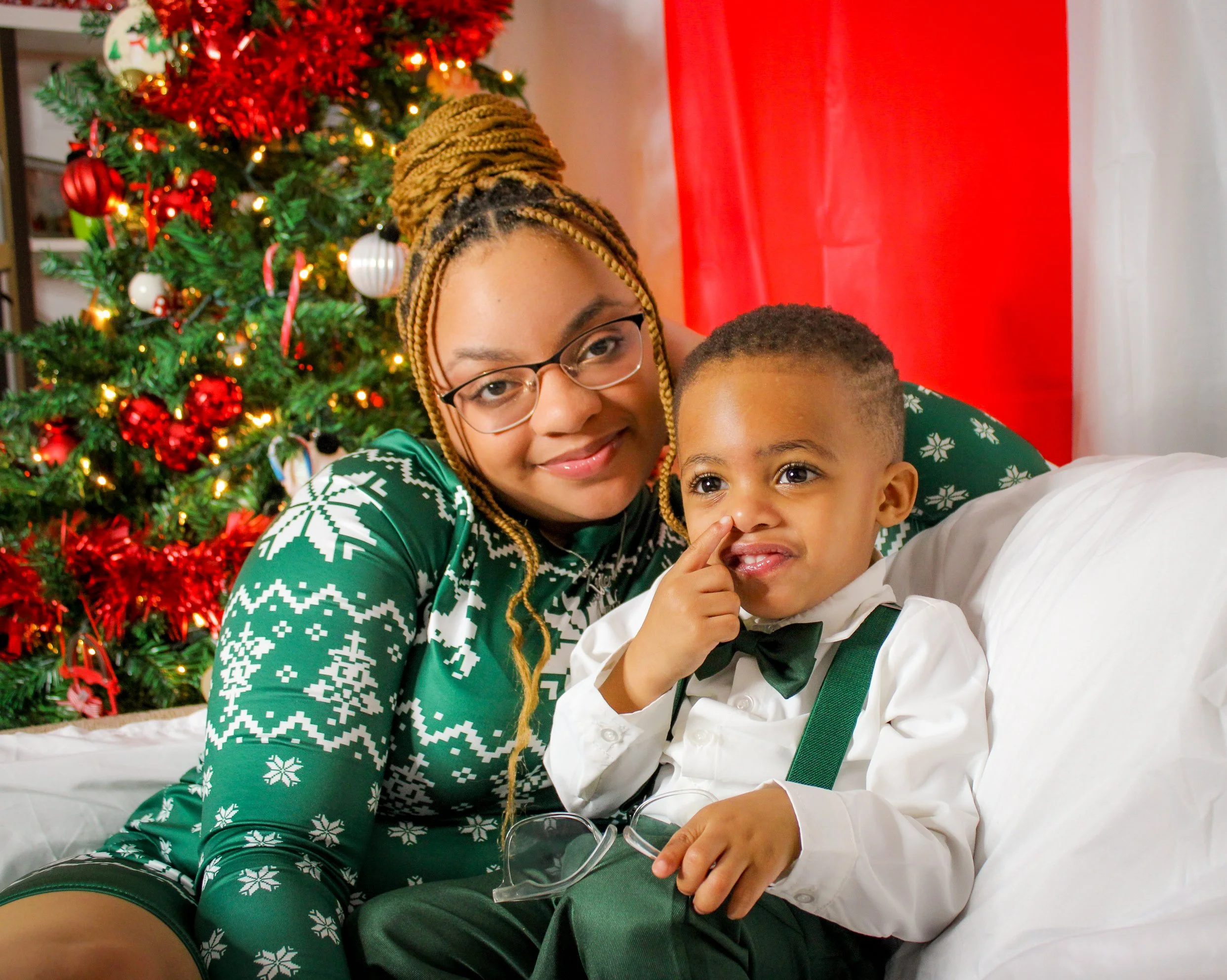 A woman with glasses and braided hair smiling, and a young boy dressed in a white shirt, black bow tie, and suspenders, sitting on a white couch in front of a decorated Christmas tree with red and gold ornaments.