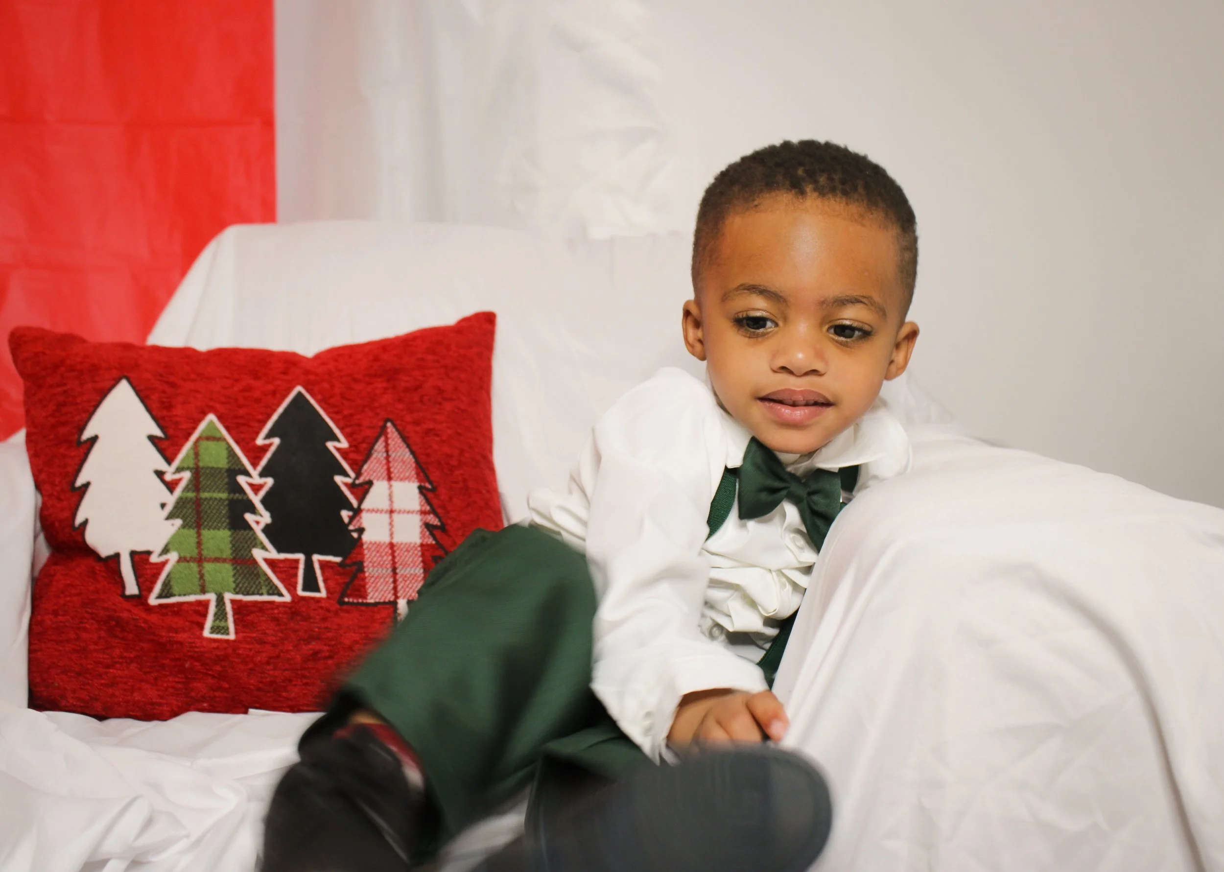 A young boy in a white shirt, black bow tie, and green pants sitting on a white couch with a red holiday pillow decorated with Christmas trees.