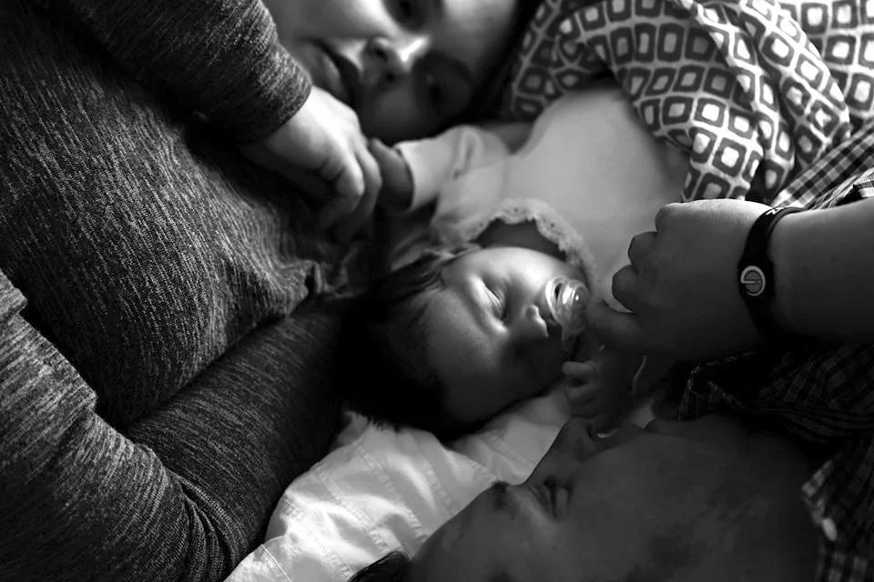 A black and white photo of two adults and two children lying on a bed, with one child in the middle having a pacifier and the other children close by.