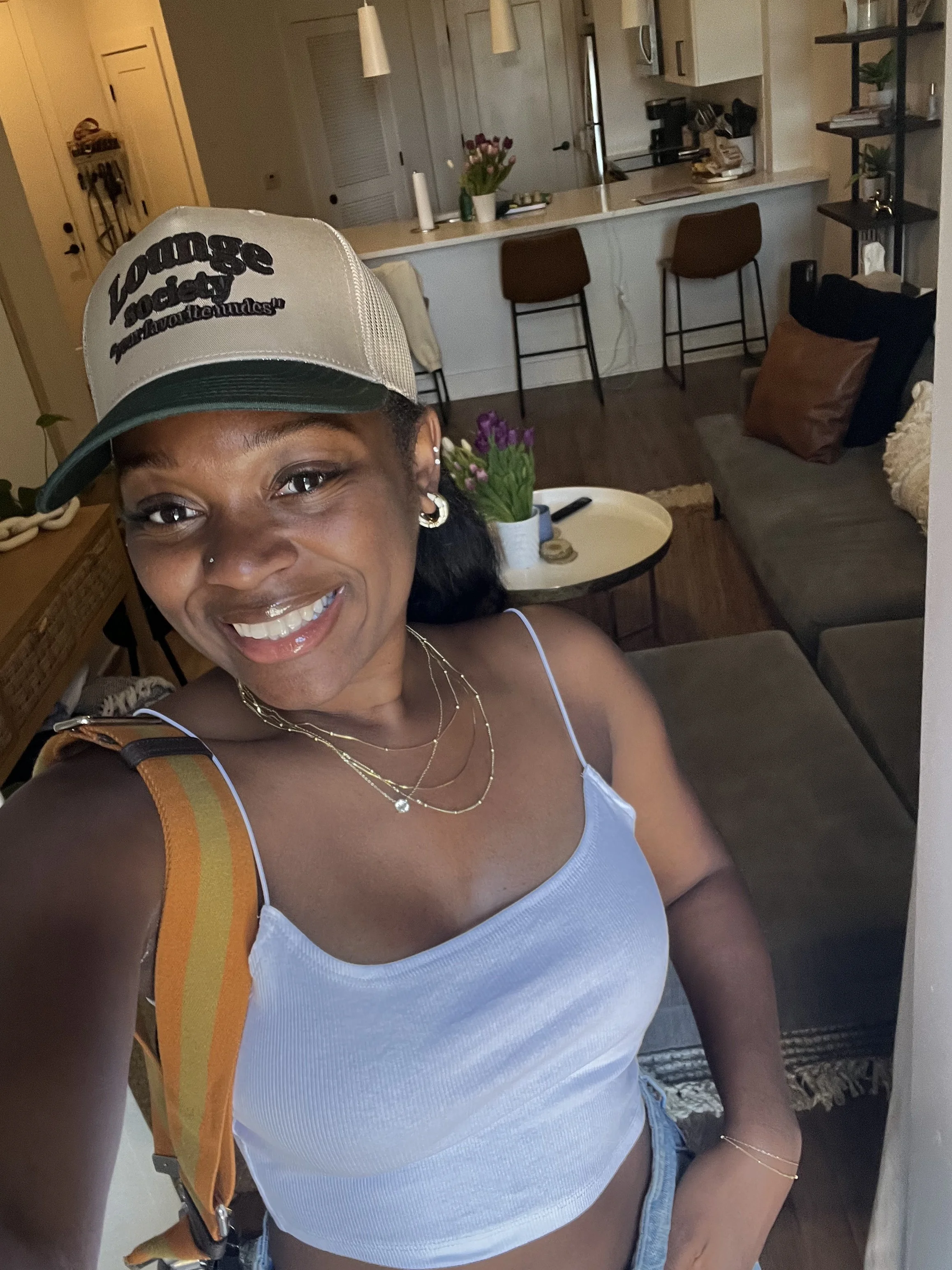 A woman smiling and taking a selfie inside a modern living room. She is wearing a beige trucker hat with black text, a white crop top, layered necklaces, and has small earrings and a nose piercing.