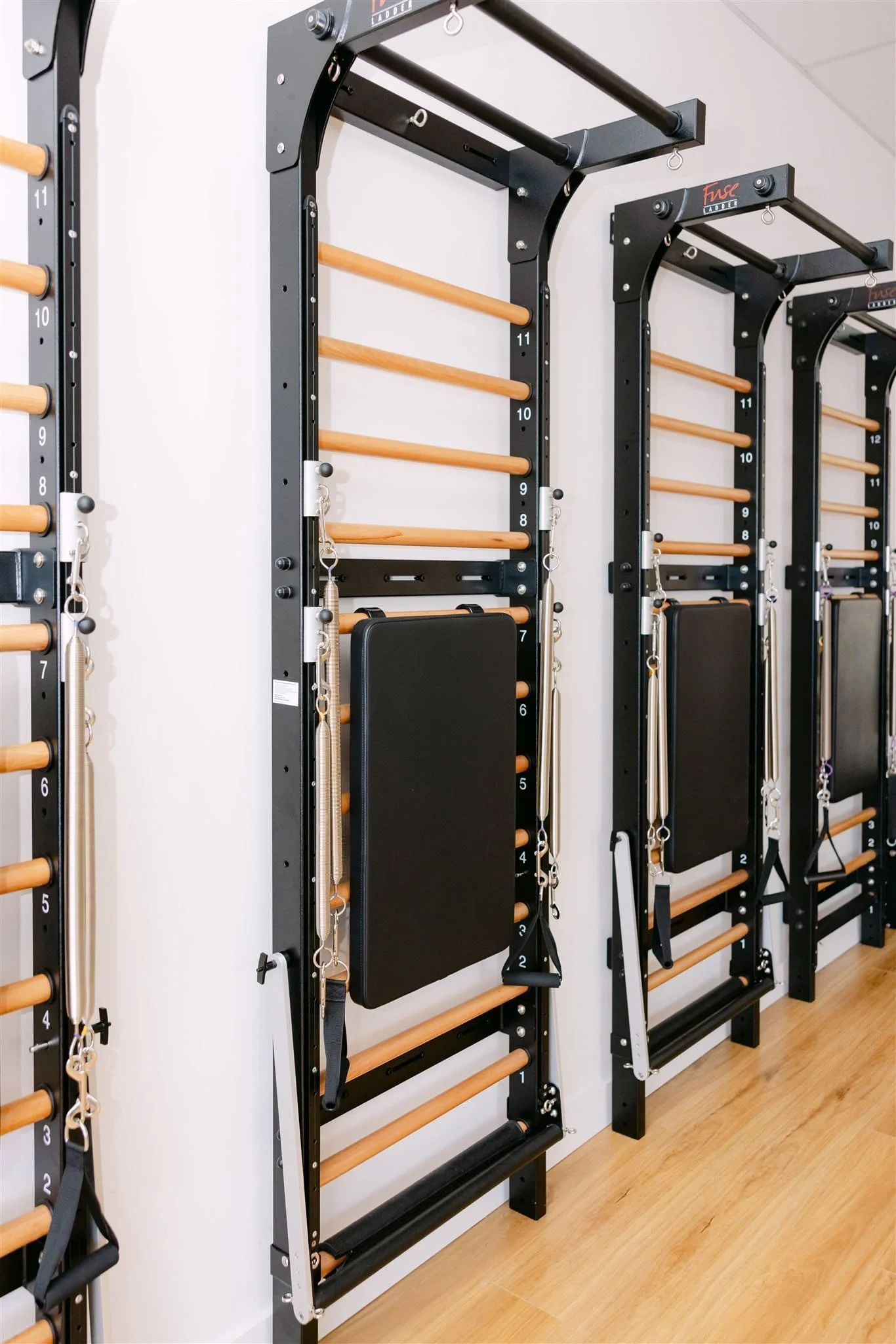 Wall of black fitness stations with wooden bars, padded boards, harnesses, and straps for exercise and physical therapy, set on a wooden floor.
